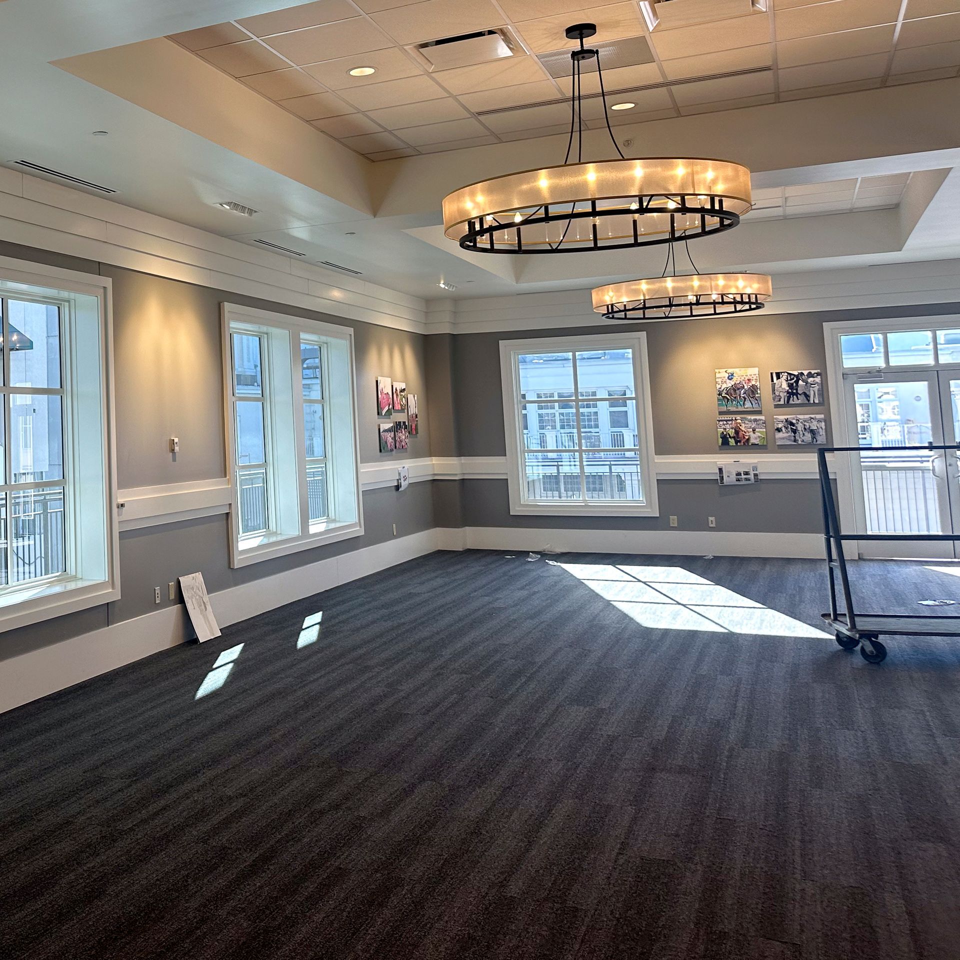 Empty room with gray walls, dark carpet, three windows, and two large chandeliers.
