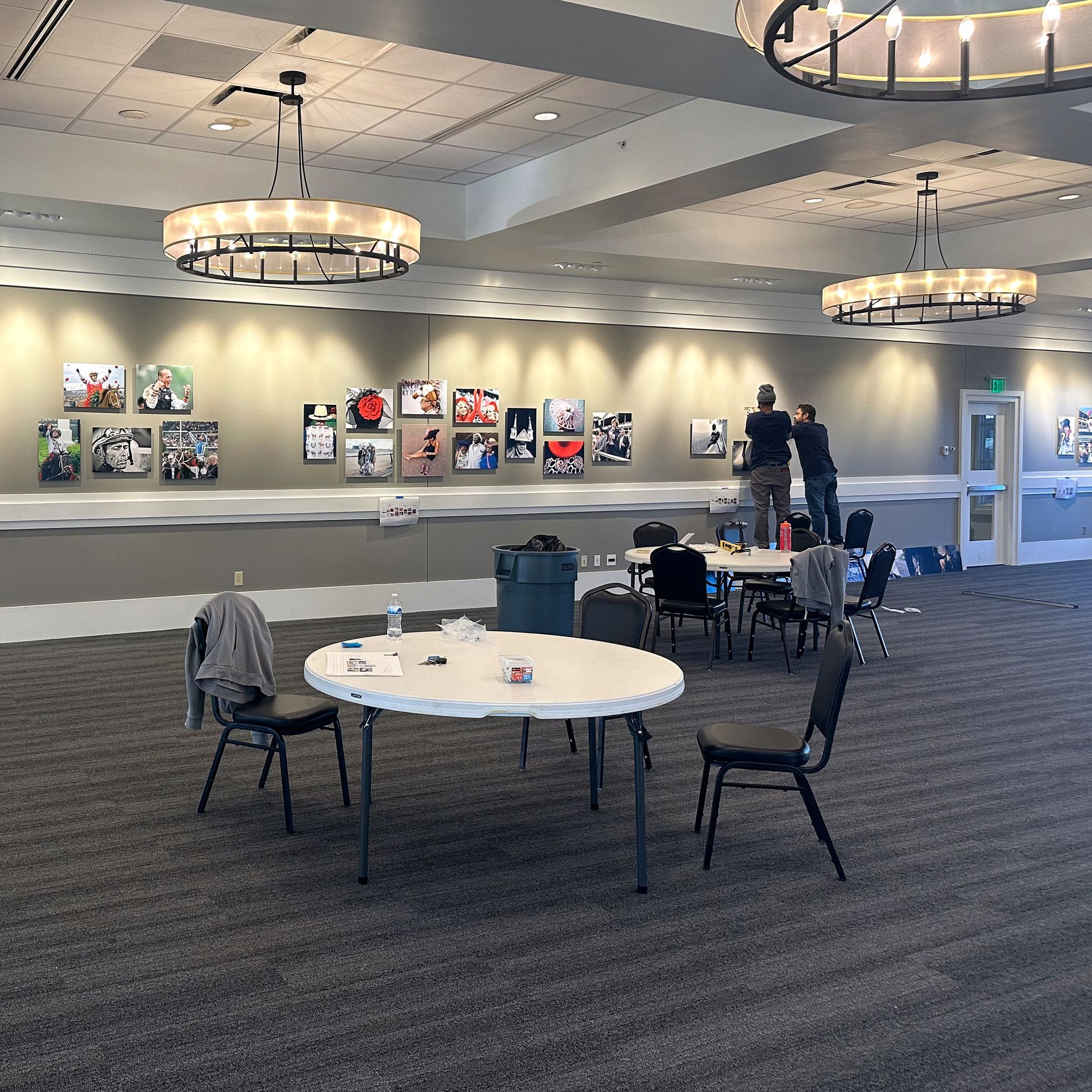 Large room with artwork on the wall, round tables, chairs, and two people adjusting the pictures.