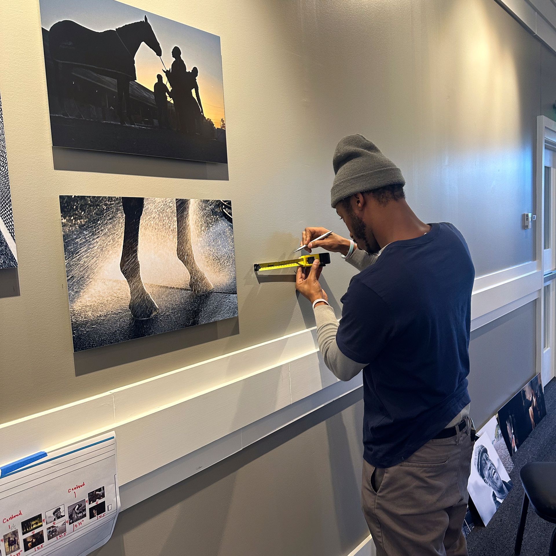 Man with beanie using a measuring tape to hang artwork on a wall. Photos of horses are already displayed.