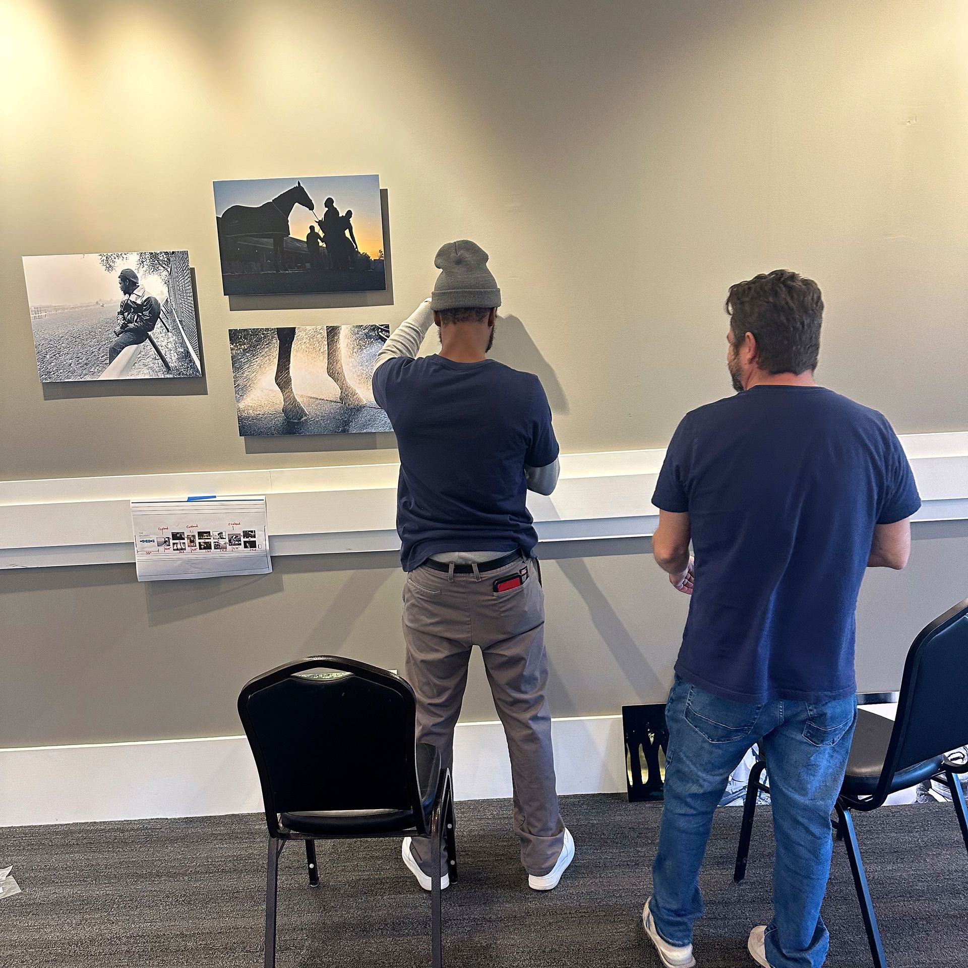Two people hanging framed photos on a wall in a room.