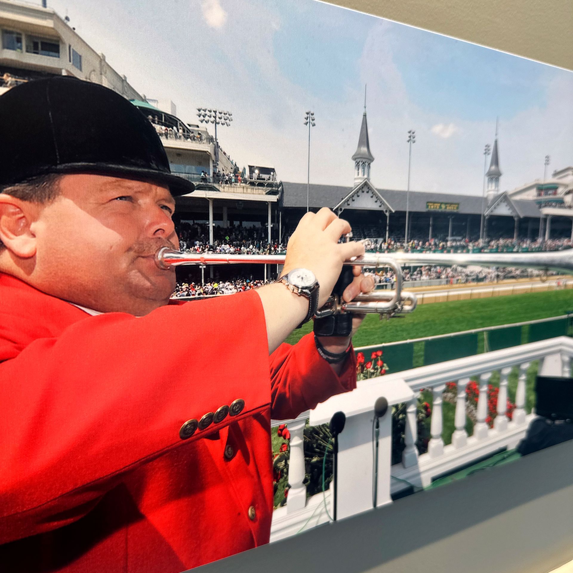 Man in red jacket and black hat playing a trumpet, with a race track in the background.