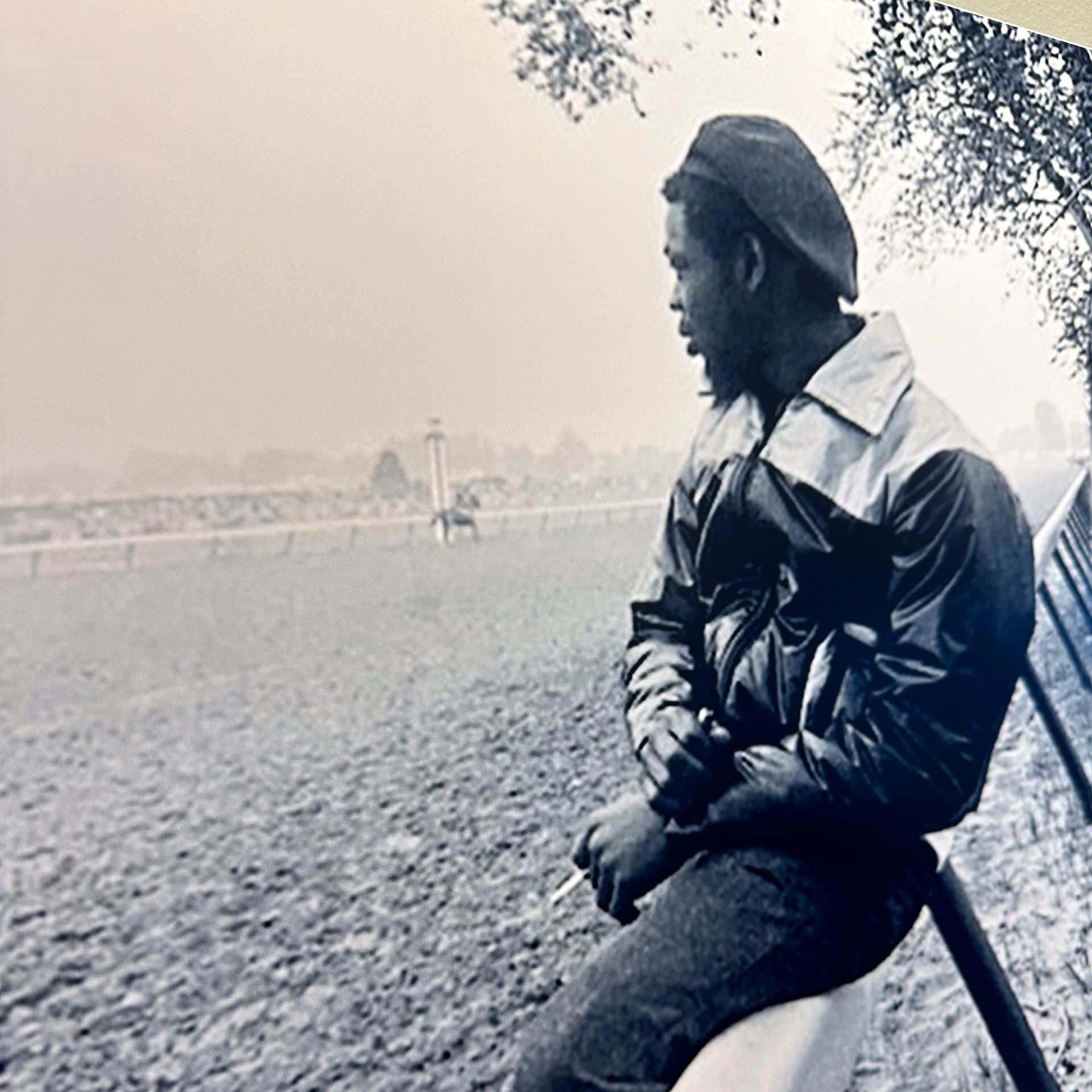 Man in jacket and cap looks at a racetrack, holding a cigarette.
