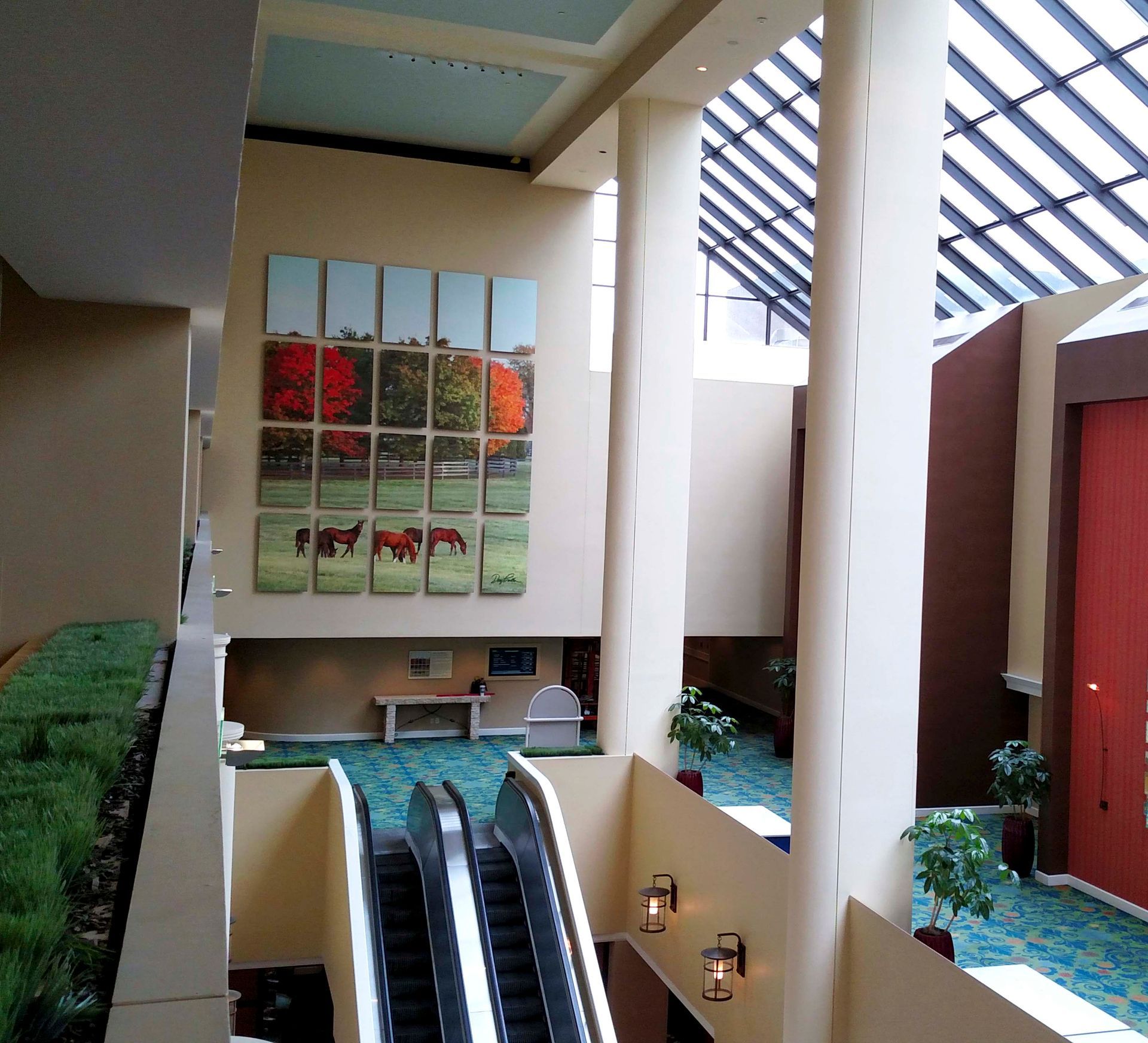 Hallway with two escalators, art, plants, and pillars. Skylight visible.