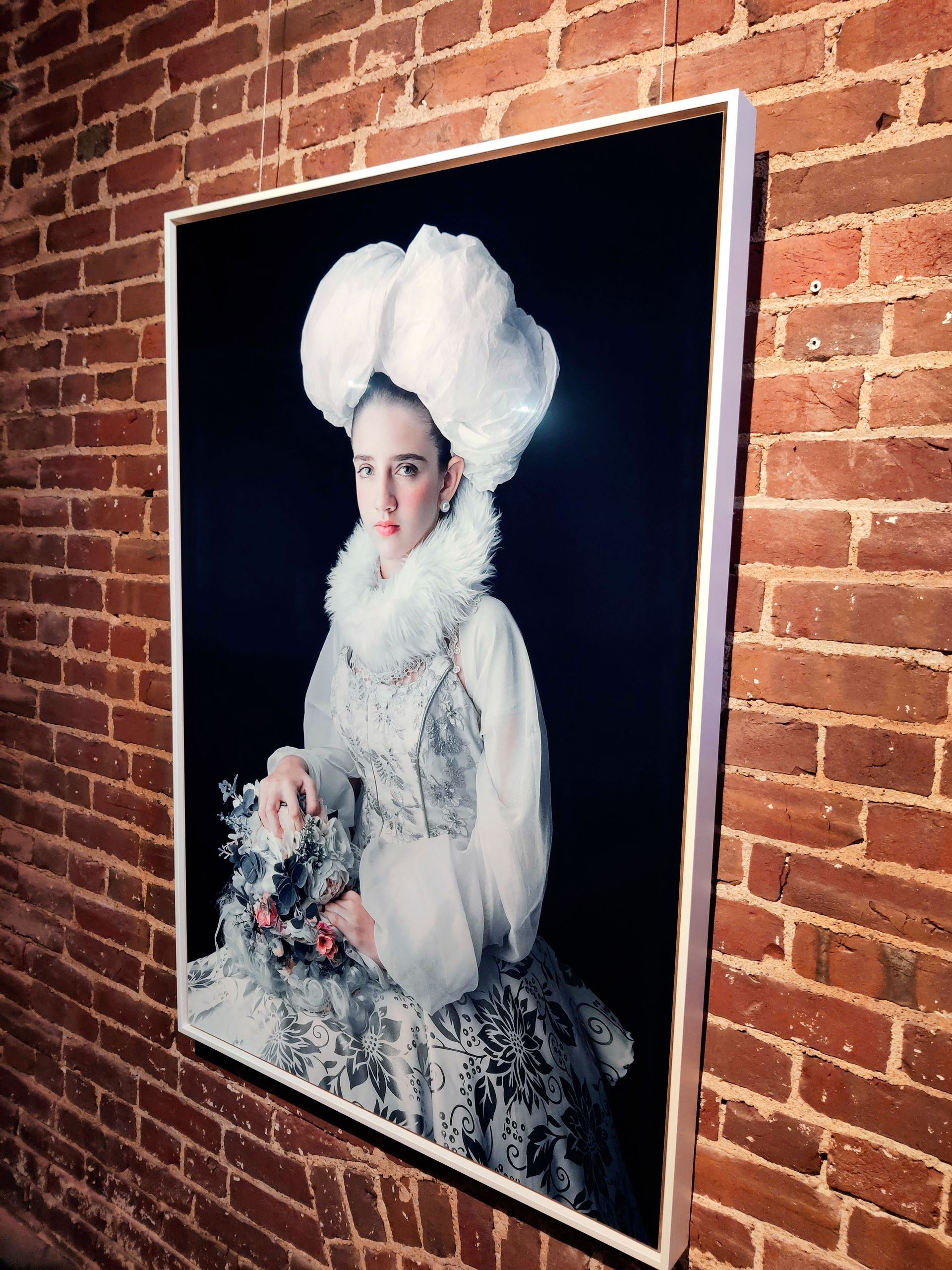 Portrait in a white frame, displayed on a brick wall. A person wearing a large white headdress, and elaborate clothing.