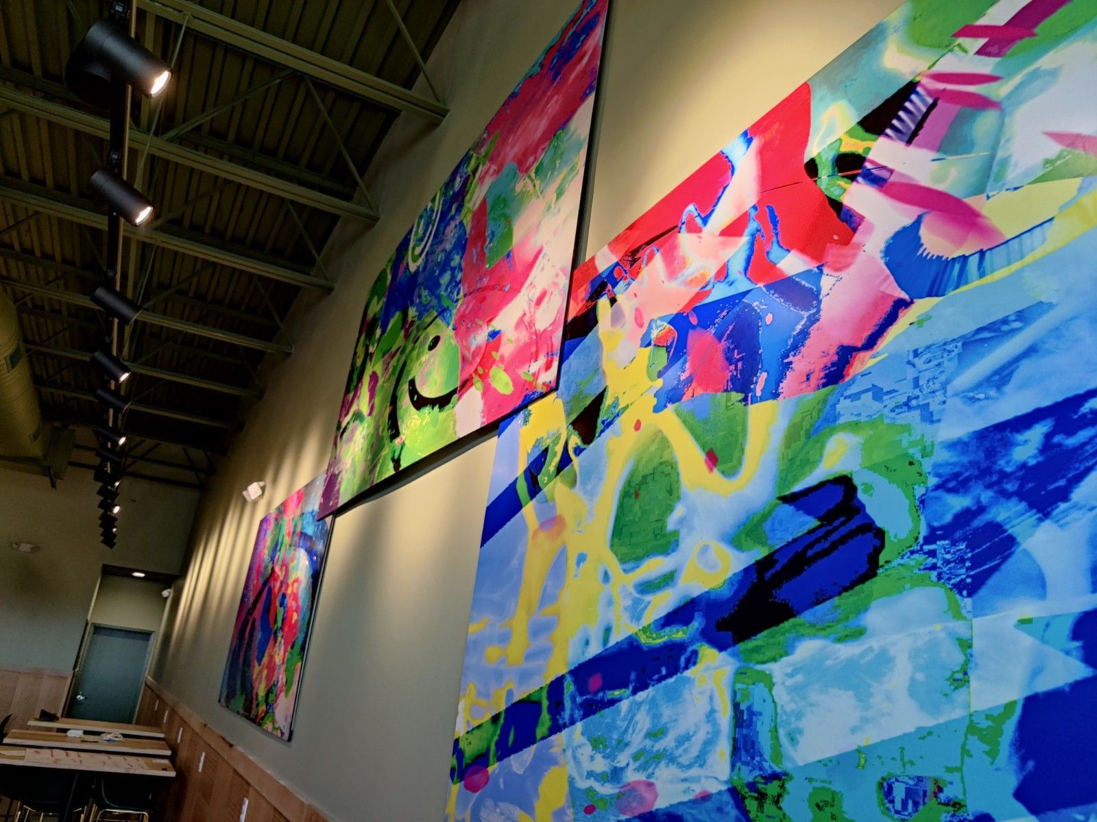Colorful abstract paintings hang on a light-colored wall, lit by spotlights, in a building.