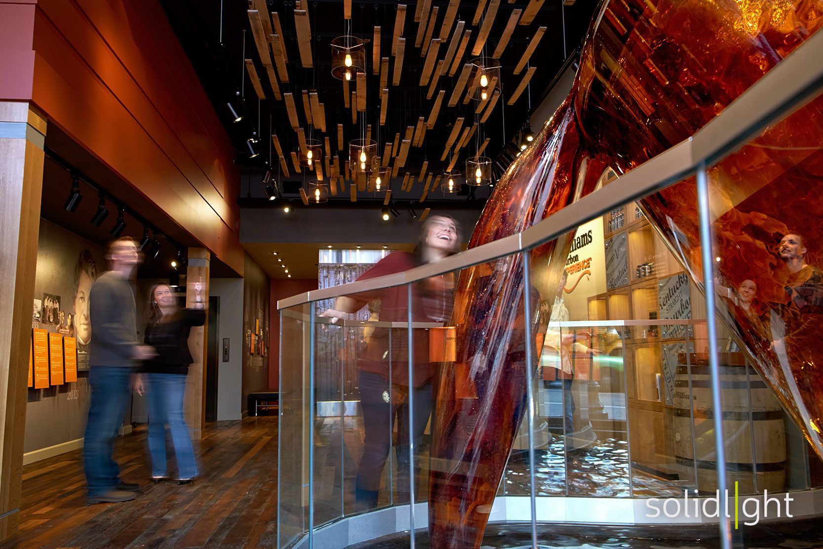 People in a museum hallway with exhibits. Glass railing in foreground, dark ceiling with lights.