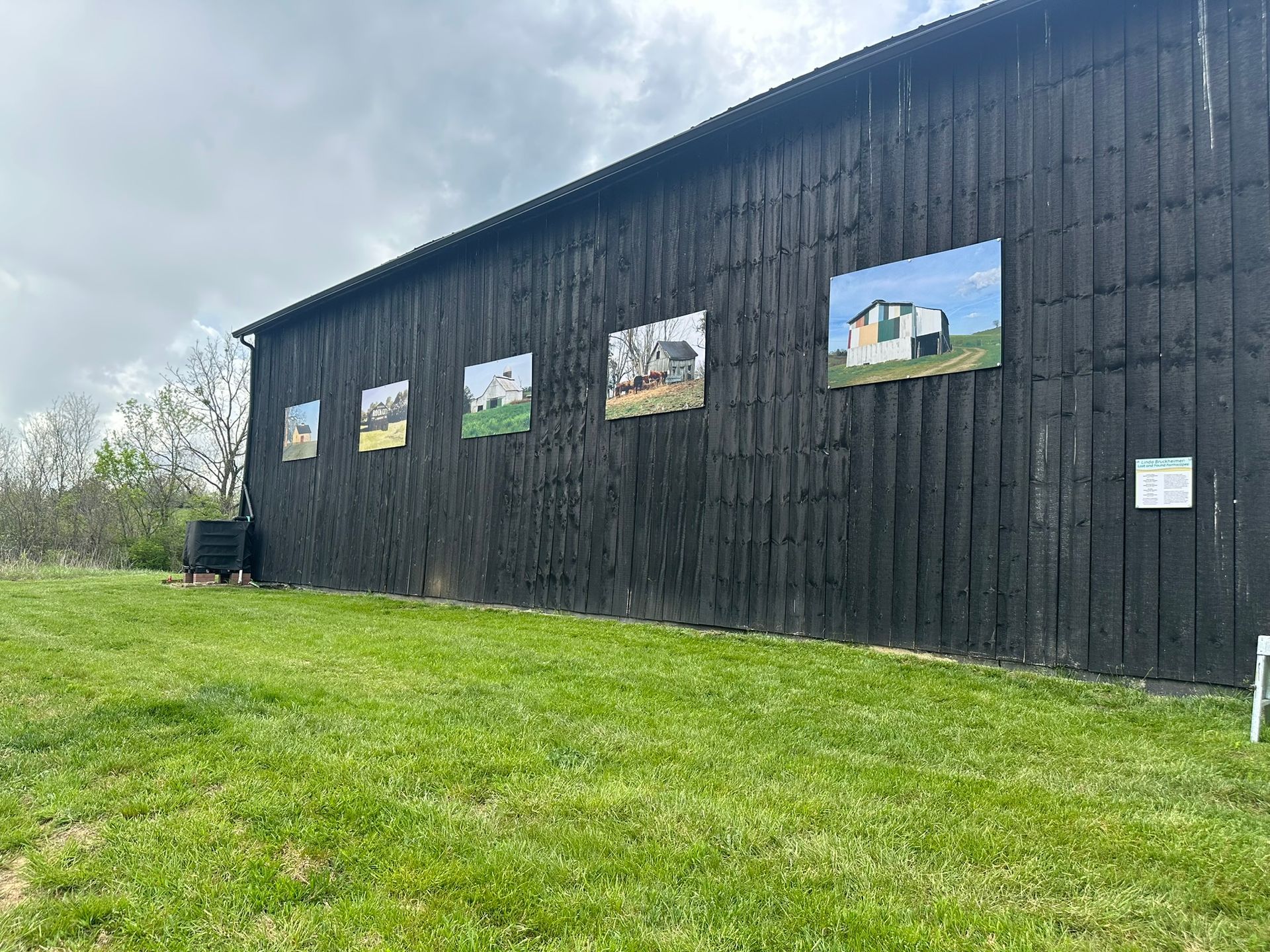 Photographs displayed on the black exterior of a building, on a grassy field, under a cloudy sky.