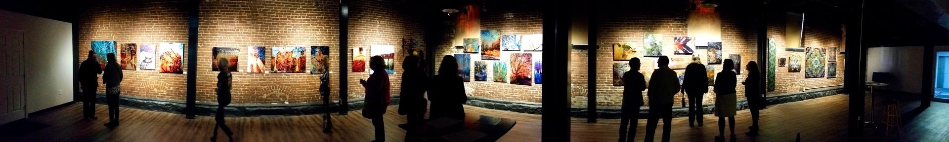 People viewing art in a gallery. Silhouetted figures stand before lit paintings on a brick wall.