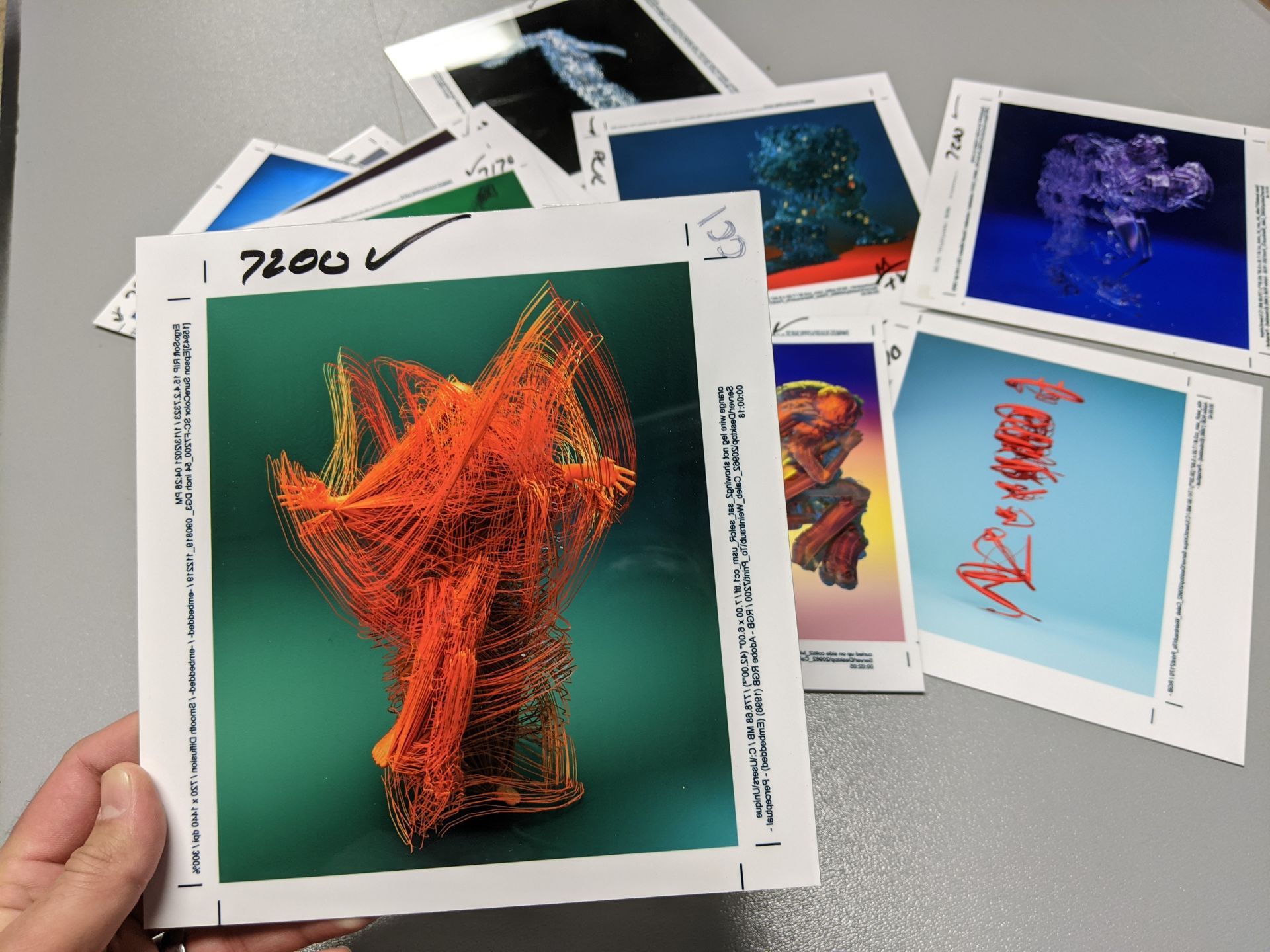 A hand holds a print with orange abstract art, surrounded by similar prints on a gray surface.