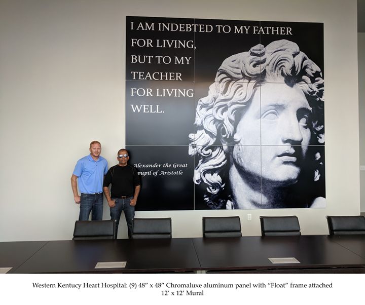 Two men standing next to a large wall panel featuring a quote and a bust of Alexander the Great.