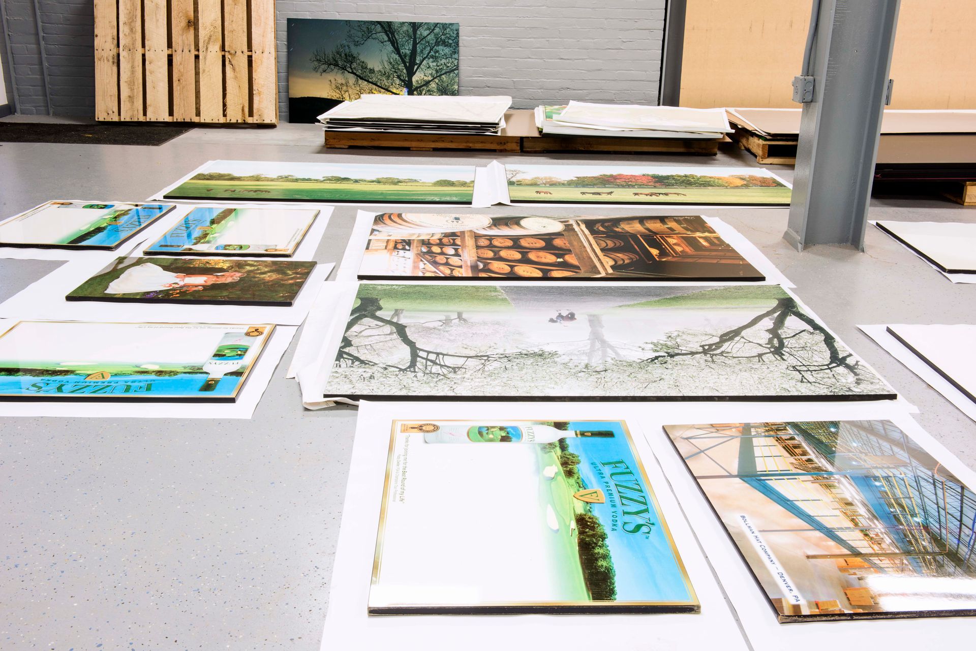 Various printed photos and artwork laid out on a gray floor in an industrial setting. Some are on pallets.