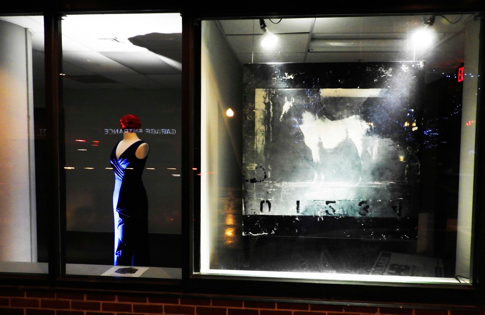 Nighttime storefront window: a blue gown on a mannequin, a large abstract artwork, reflections of city lights.
