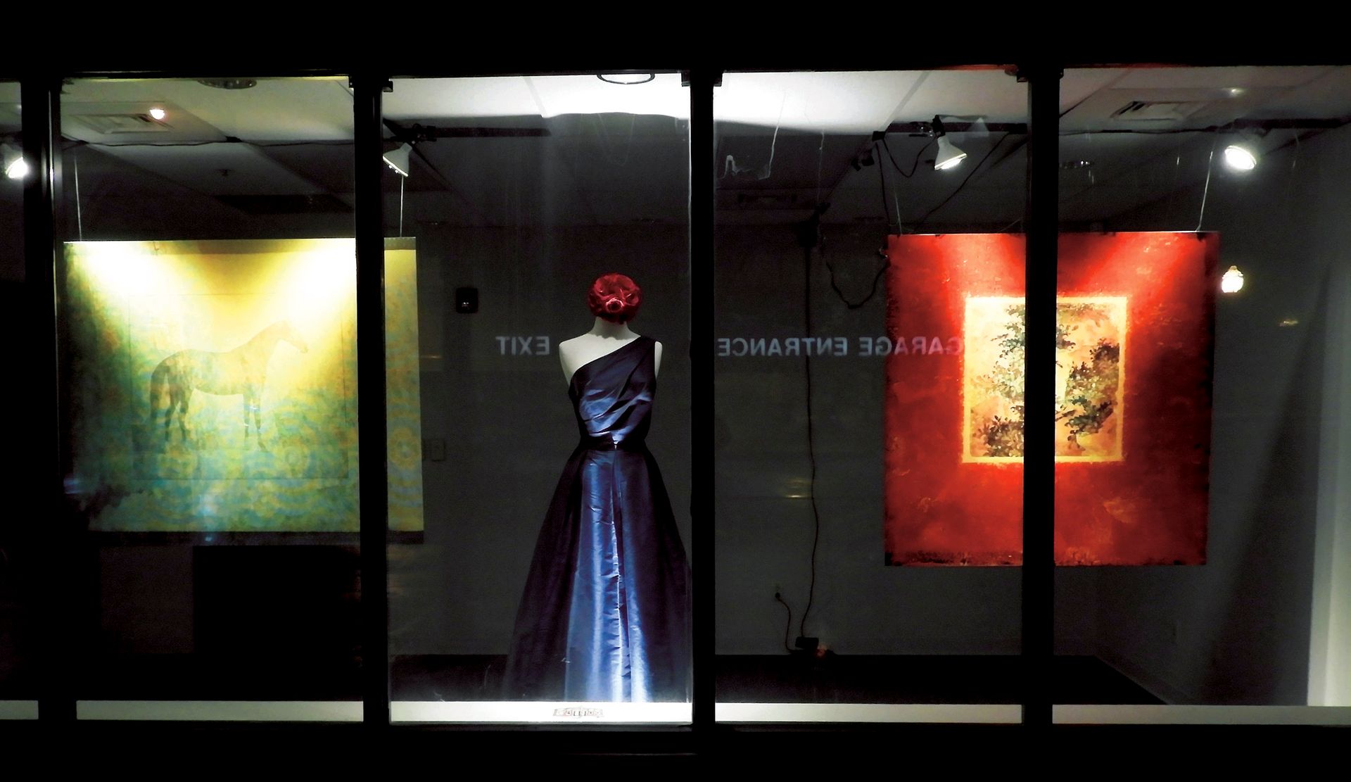 Storefront window display with a blue dress on a mannequin between two illuminated artworks.