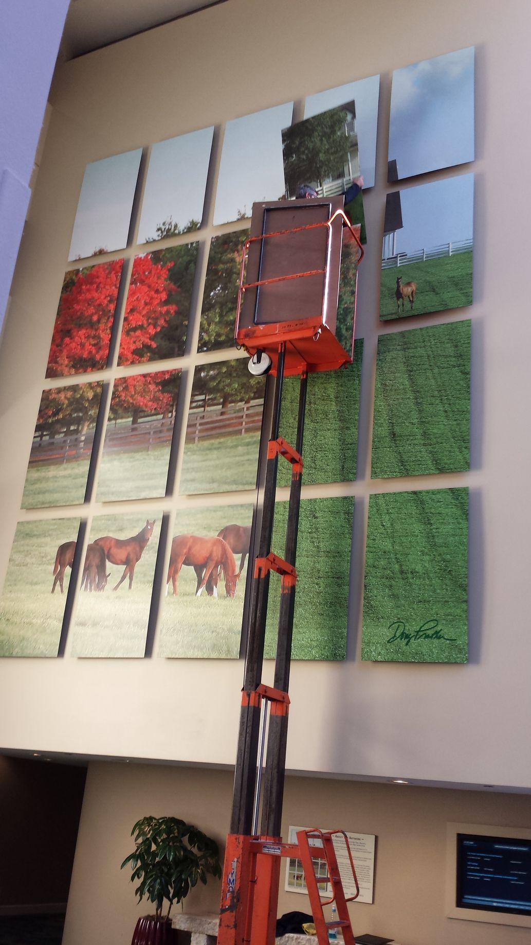 An orange lift truck near a wall with large framed art panels featuring landscapes and horses.