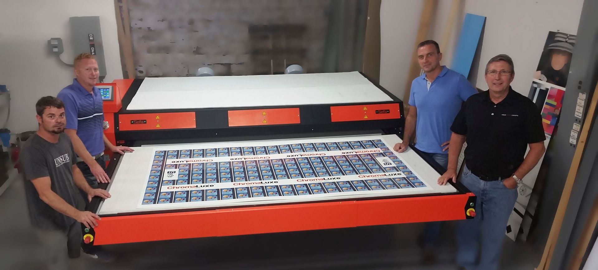 Four men standing around a large orange machine printing a design in a workshop.