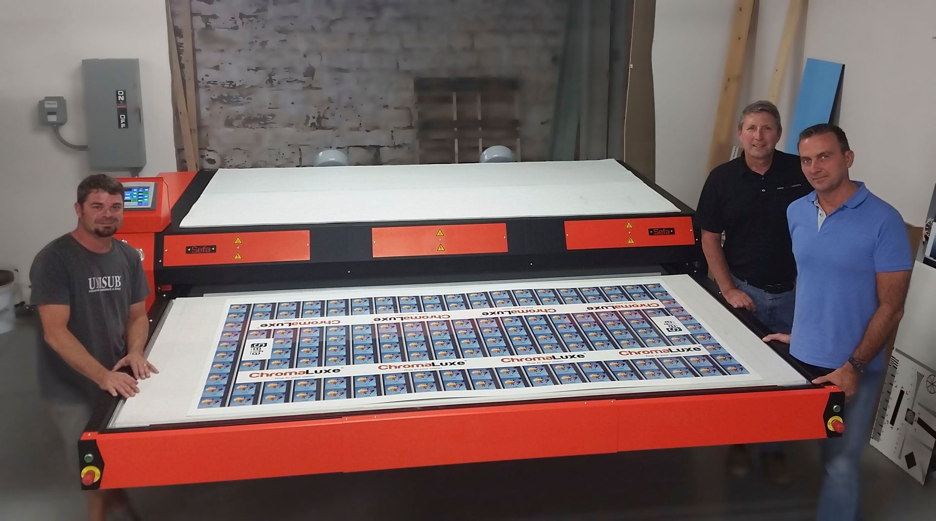 Three people holding a large printed graphic at an orange printing press in a shop.