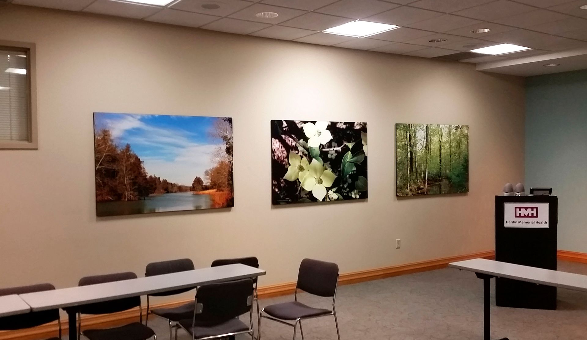 Three landscape photos hang on a beige wall in a room with a podium and chairs.