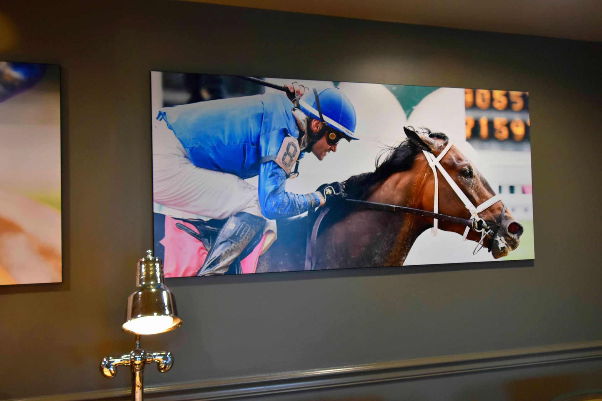 Jockey in blue racing silks on a brown horse, close-up, against white backdrop; framed on gray wall.