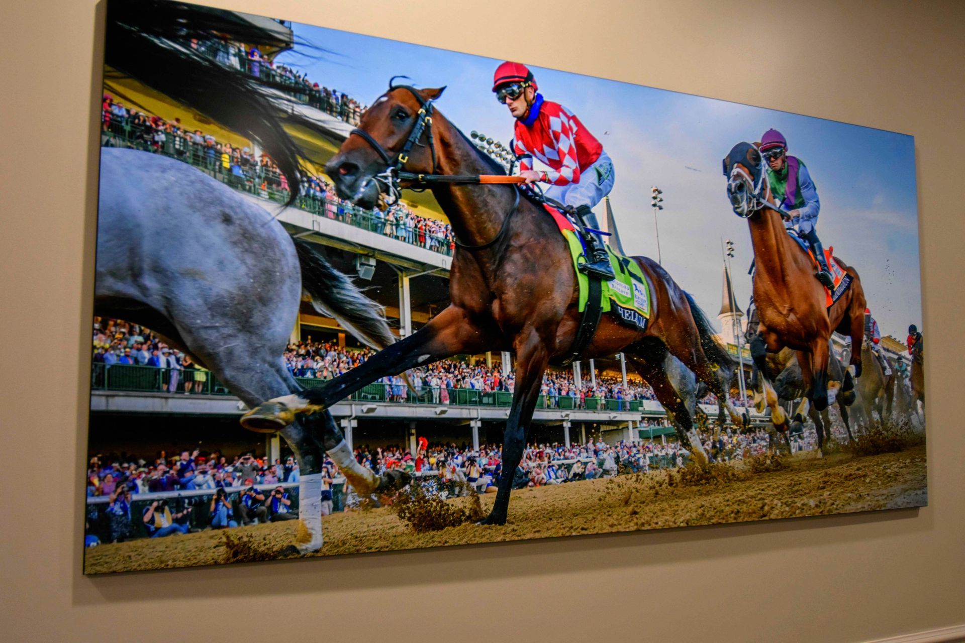Racehorses rounding a turn on a dirt track, jockeys in colorful silks. Fans in the stands.