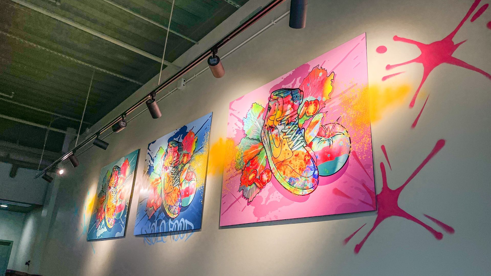 Three colorful abstract paintings on a wall with paint splatters. Spotlights shine above.