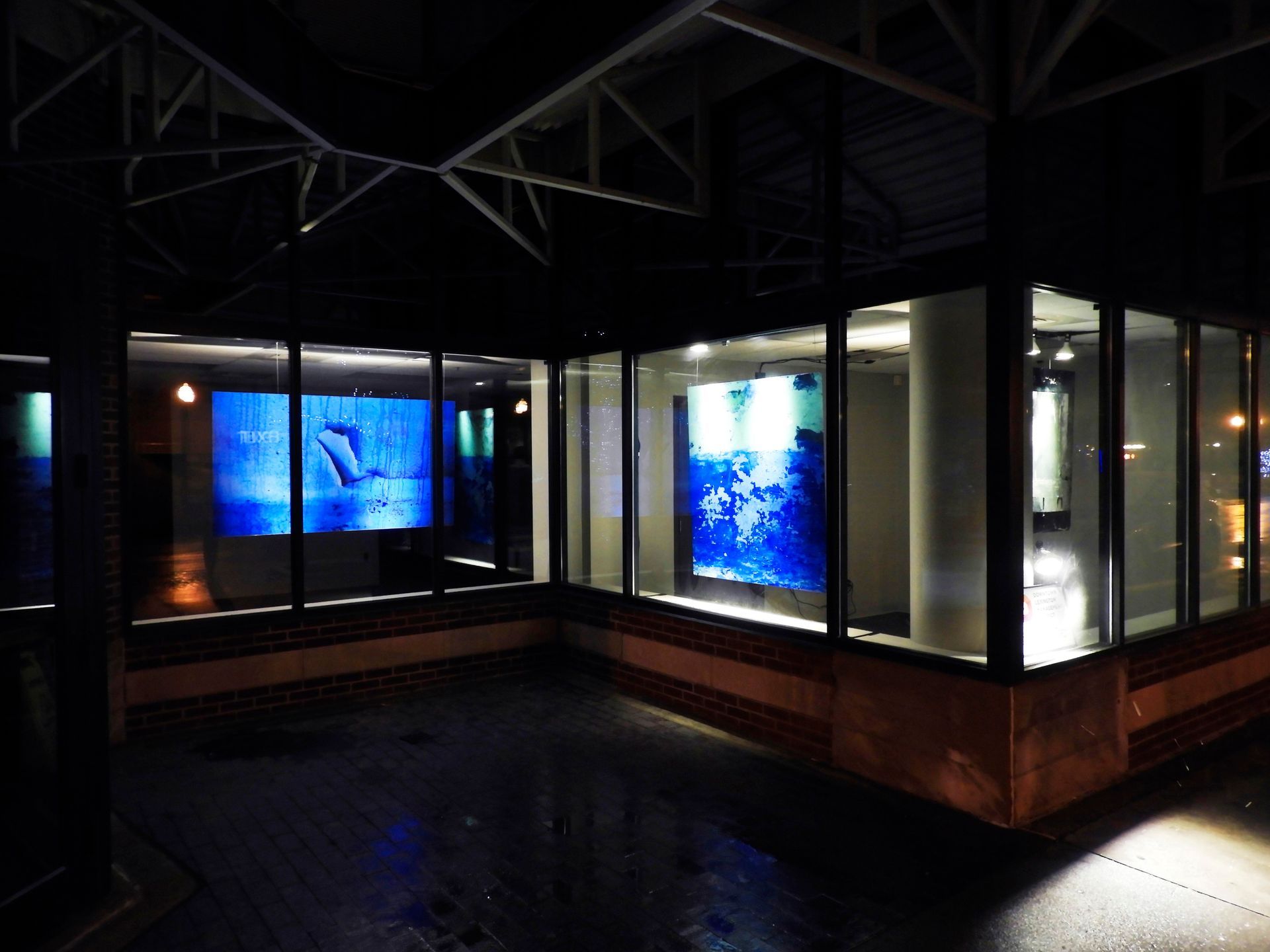 Glass-enclosed gallery displays two blue-lit art pieces at night; brick and concrete exterior with dark ceiling.