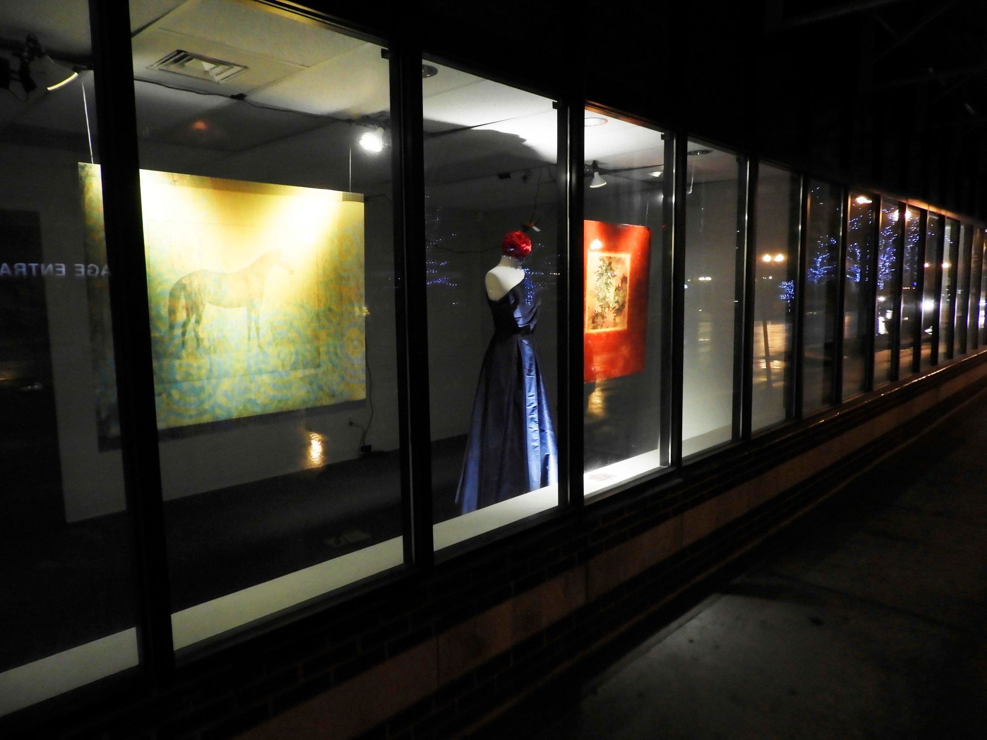 Museum window displays at night; art on walls, mannequin in blue dress, lights.
