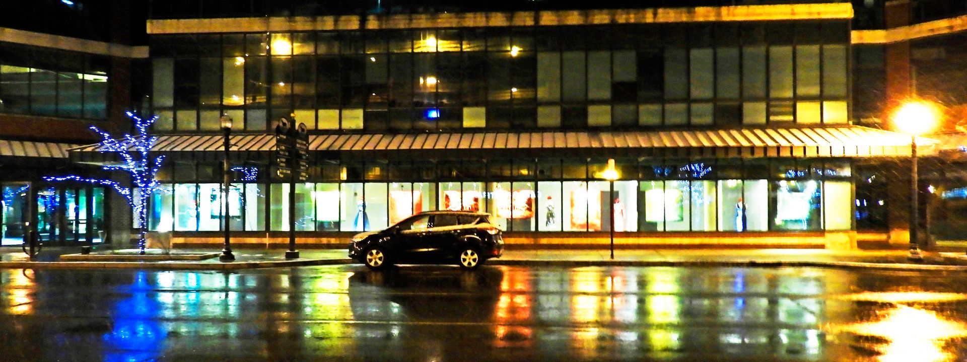 Nighttime cityscape with a modern building and a wet street reflecting lights. A car is parked out front.
