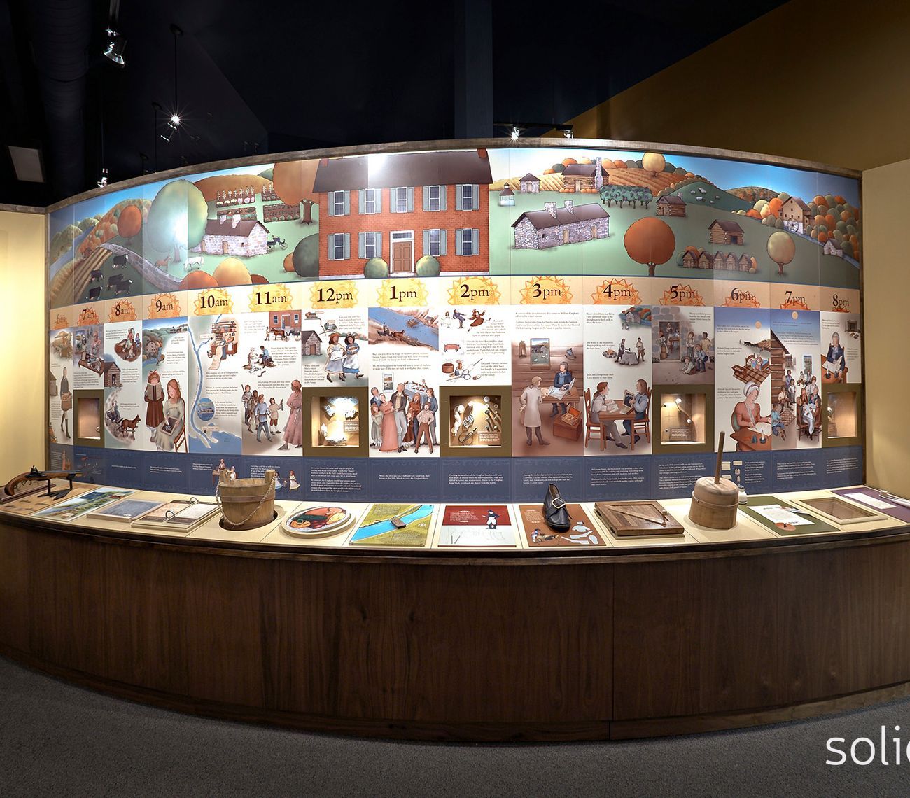 Museum display featuring a circular mural of a countryside scene above illustrations and artifacts on a curved wooden counter.