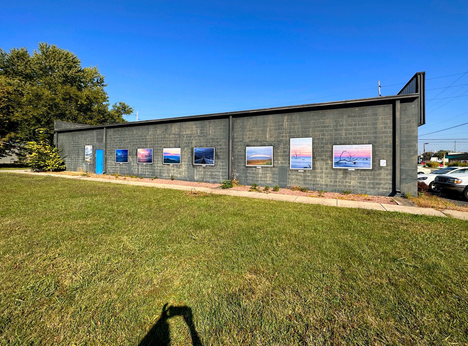 A brick building with several framed paintings on the wall; grassy lawn in the foreground; blue sky.