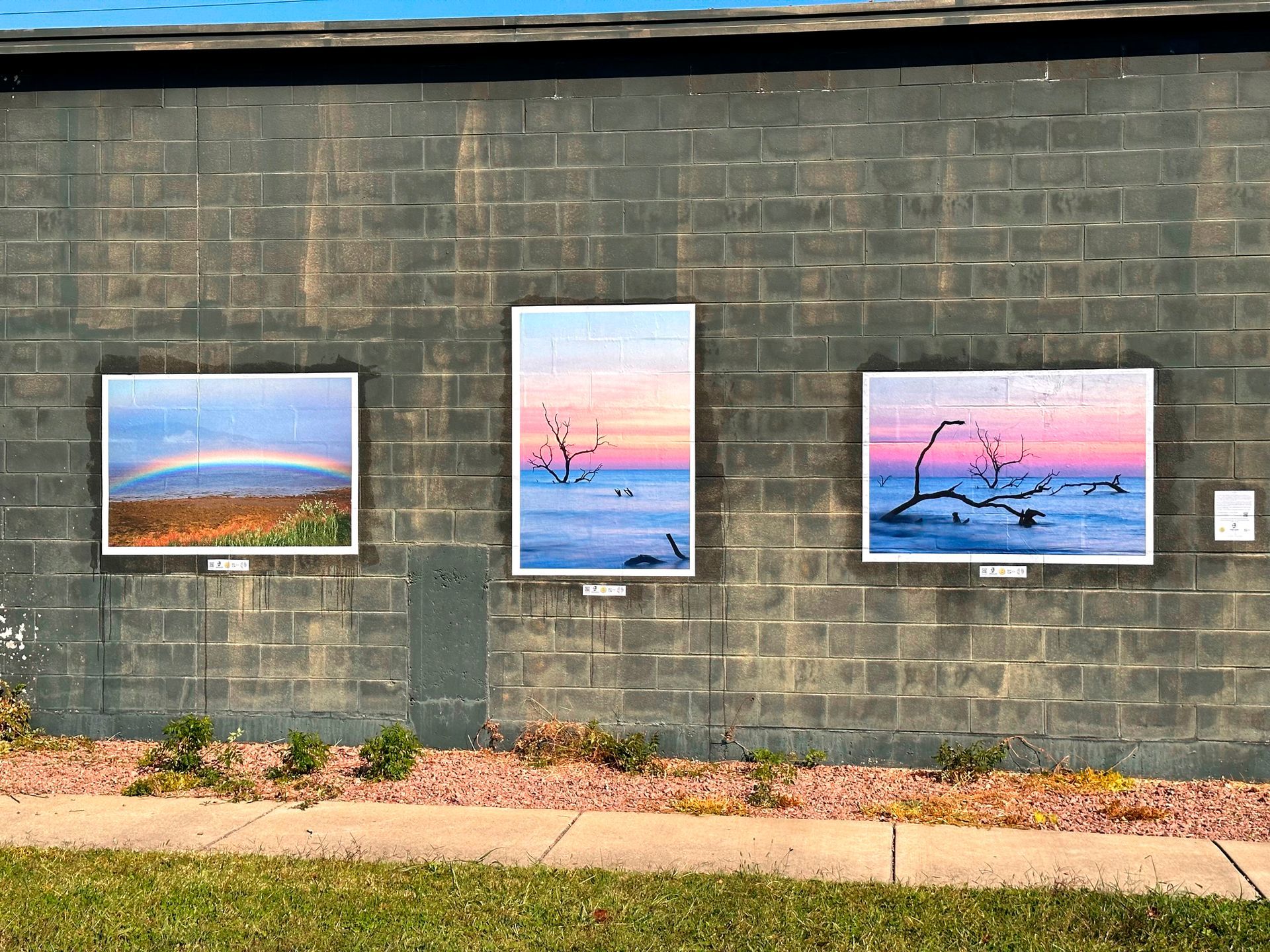 Three framed landscape puzzles on a gray brick wall: rainbow, sunrise, and sunset over water with bare branches.