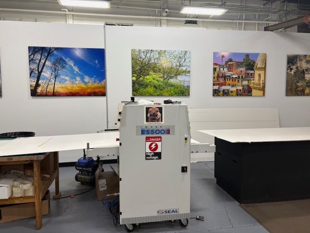 Large-format printer in a print shop, with mounted art on the wall behind it.