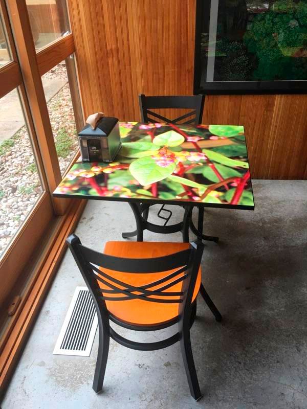Small table with two chairs in a room. Table has a colorful floral design, one chair is orange. Wooden walls, a window, and a vent.