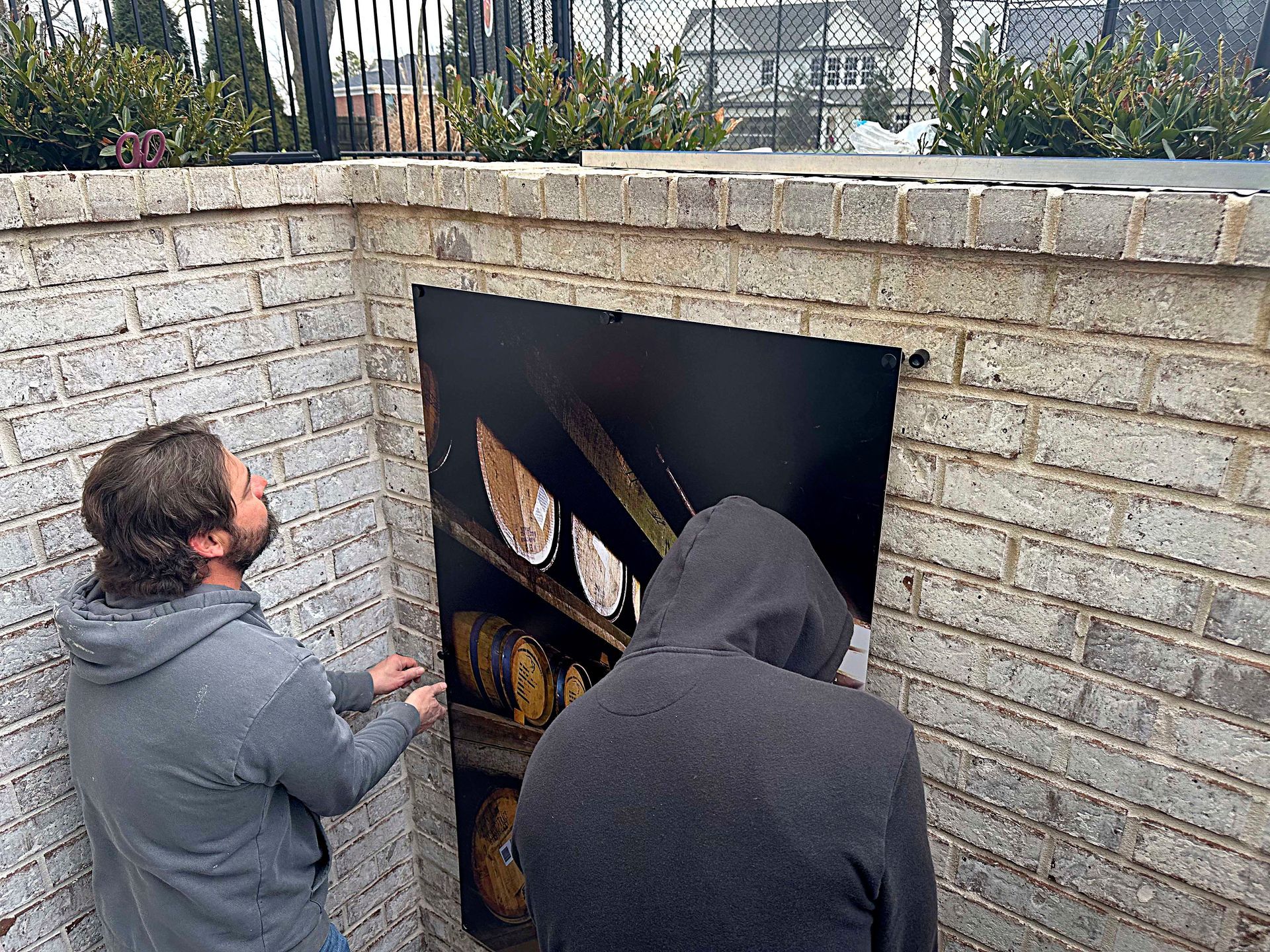 Two people installing a black panel in a brick wall. The panel has an image of wooden barrels.