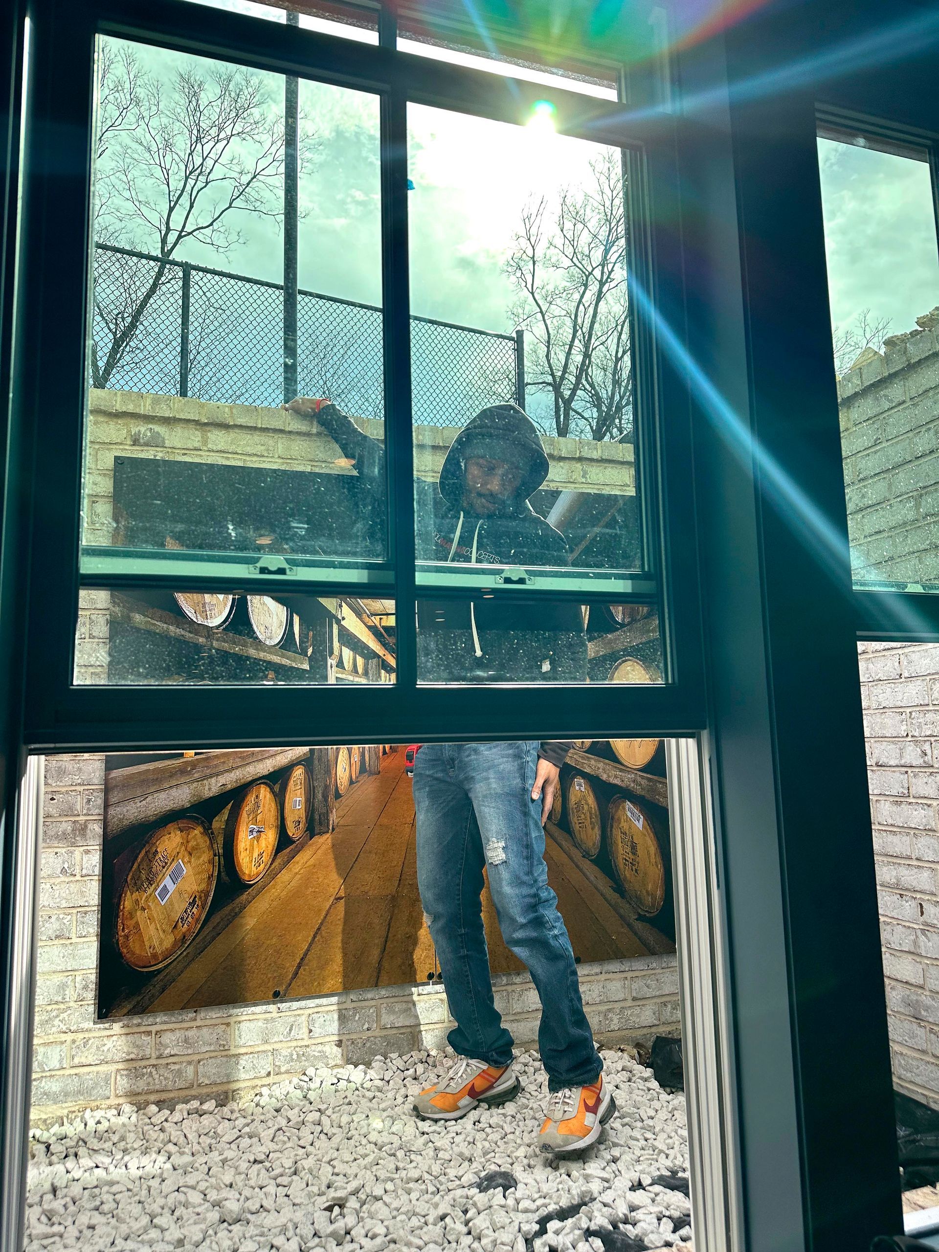 Person in jeans and orange shoes stands in front of a window, tank tracks art below, sunlight through trees above.