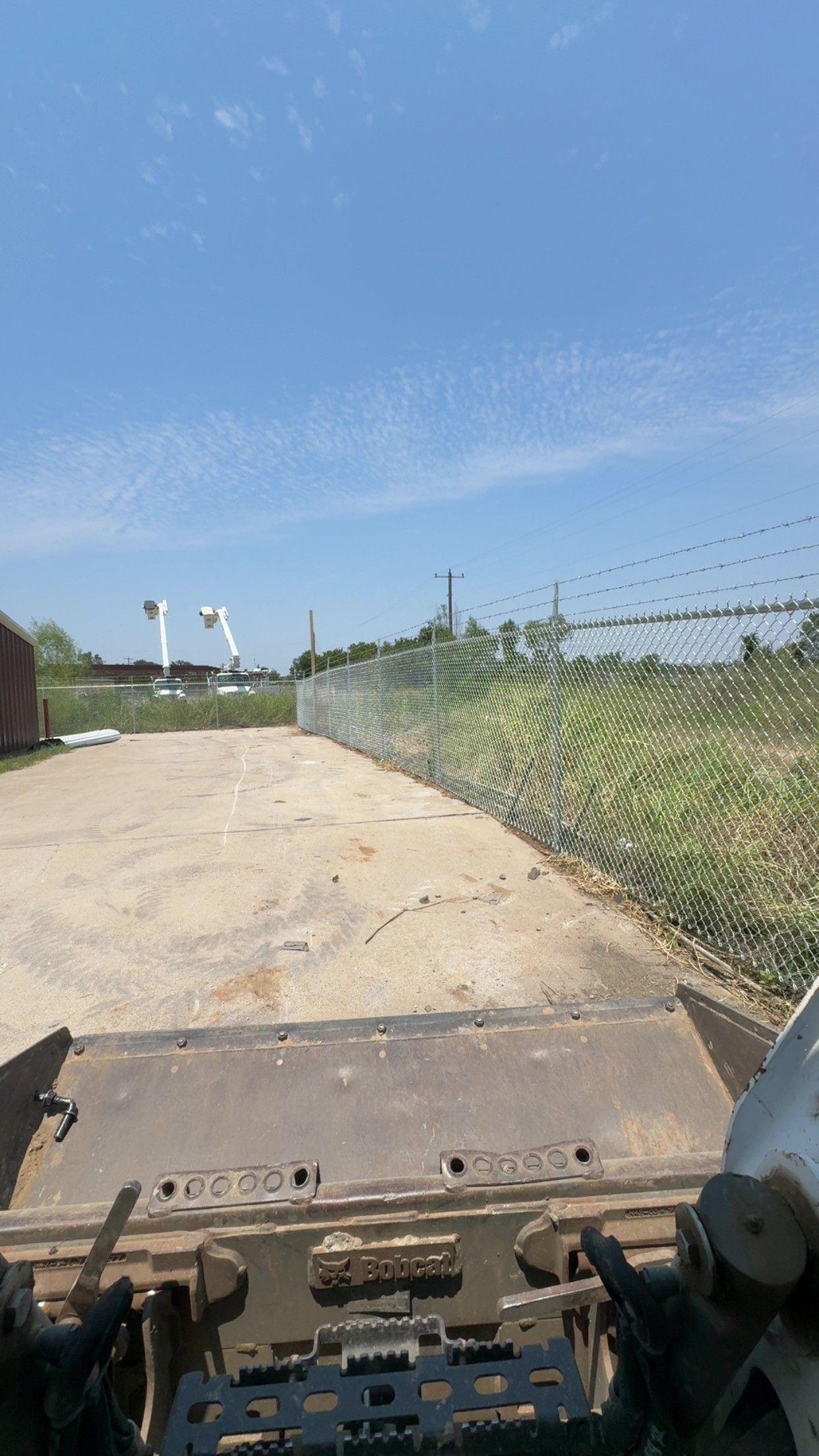 Paved path beside a chain-link fence under a clear blue sky, viewed from a vehicle or machinery.