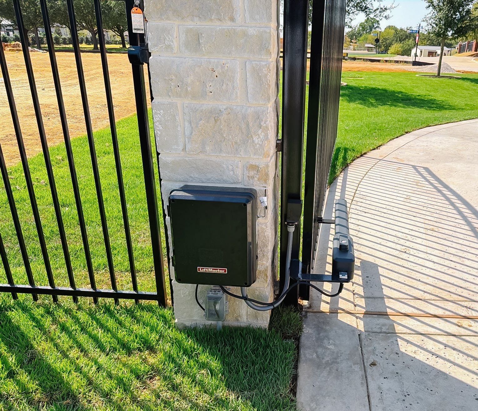 Black utility box on a stone gate pillar beside a metal fence and paved walkway.