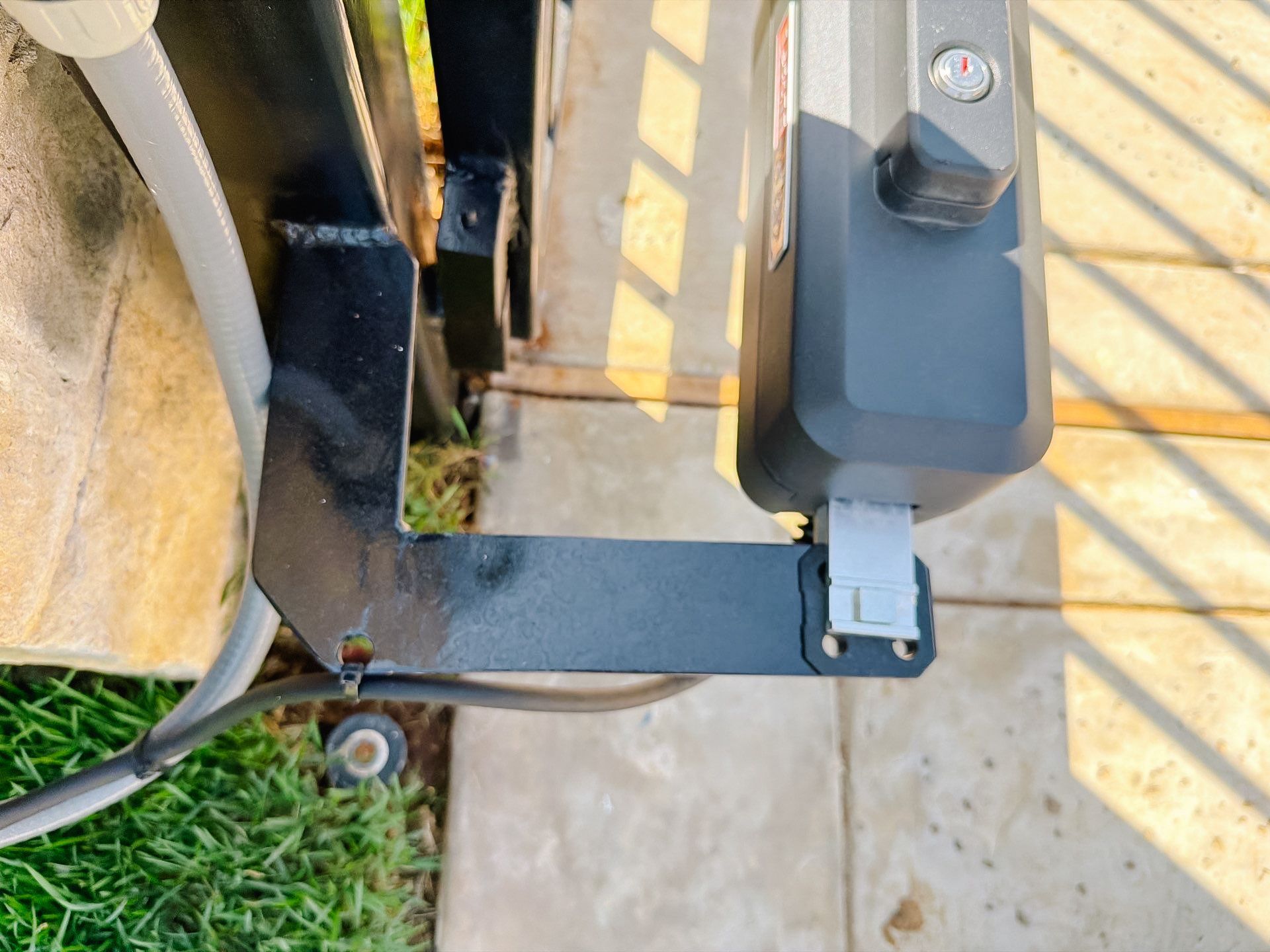 Close-up of a black metal gate hinge and latch on a concrete patio beside grass.