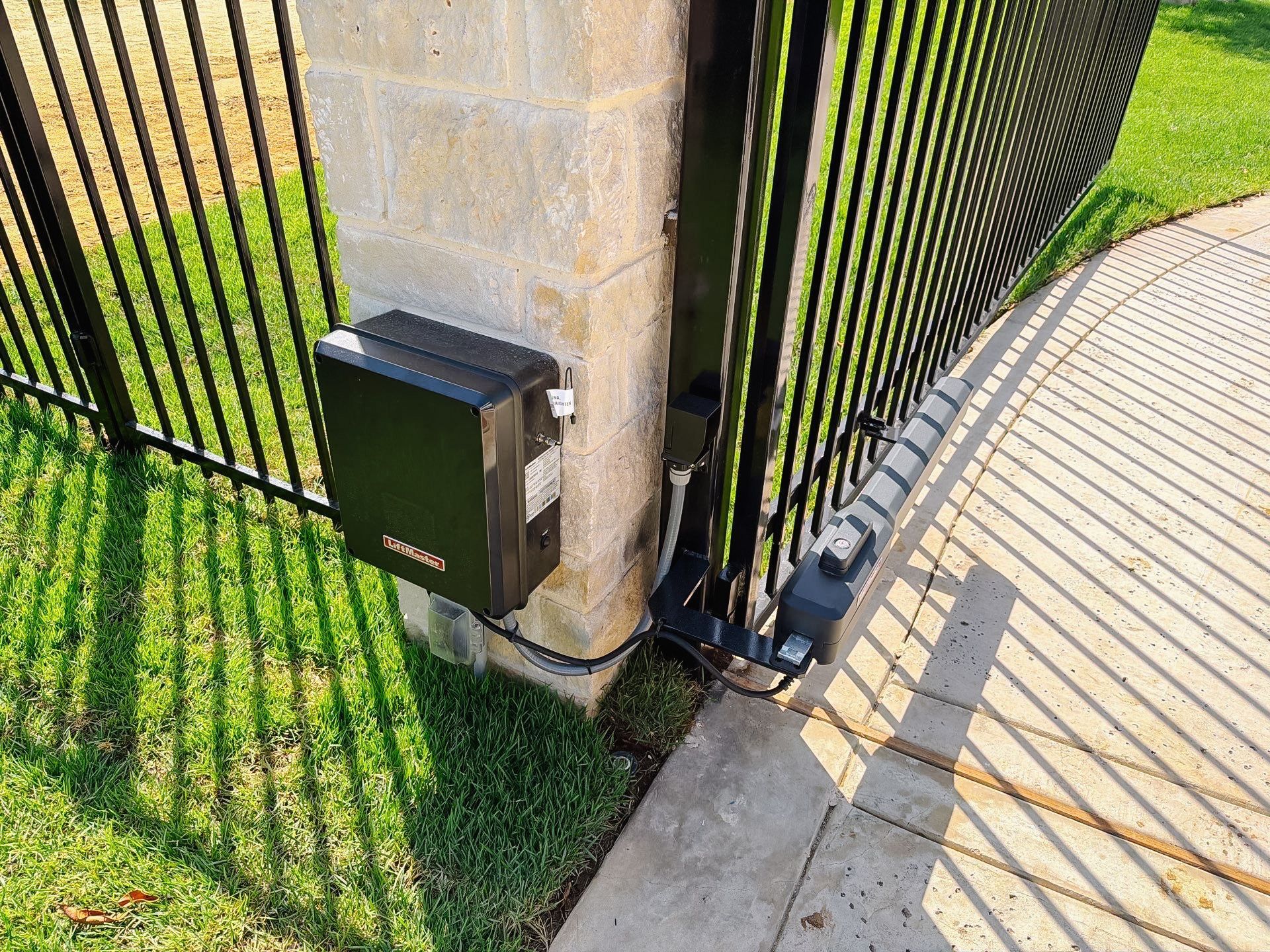 Black gate control box mounted on a stone post beside a metal fence and sidewalk.