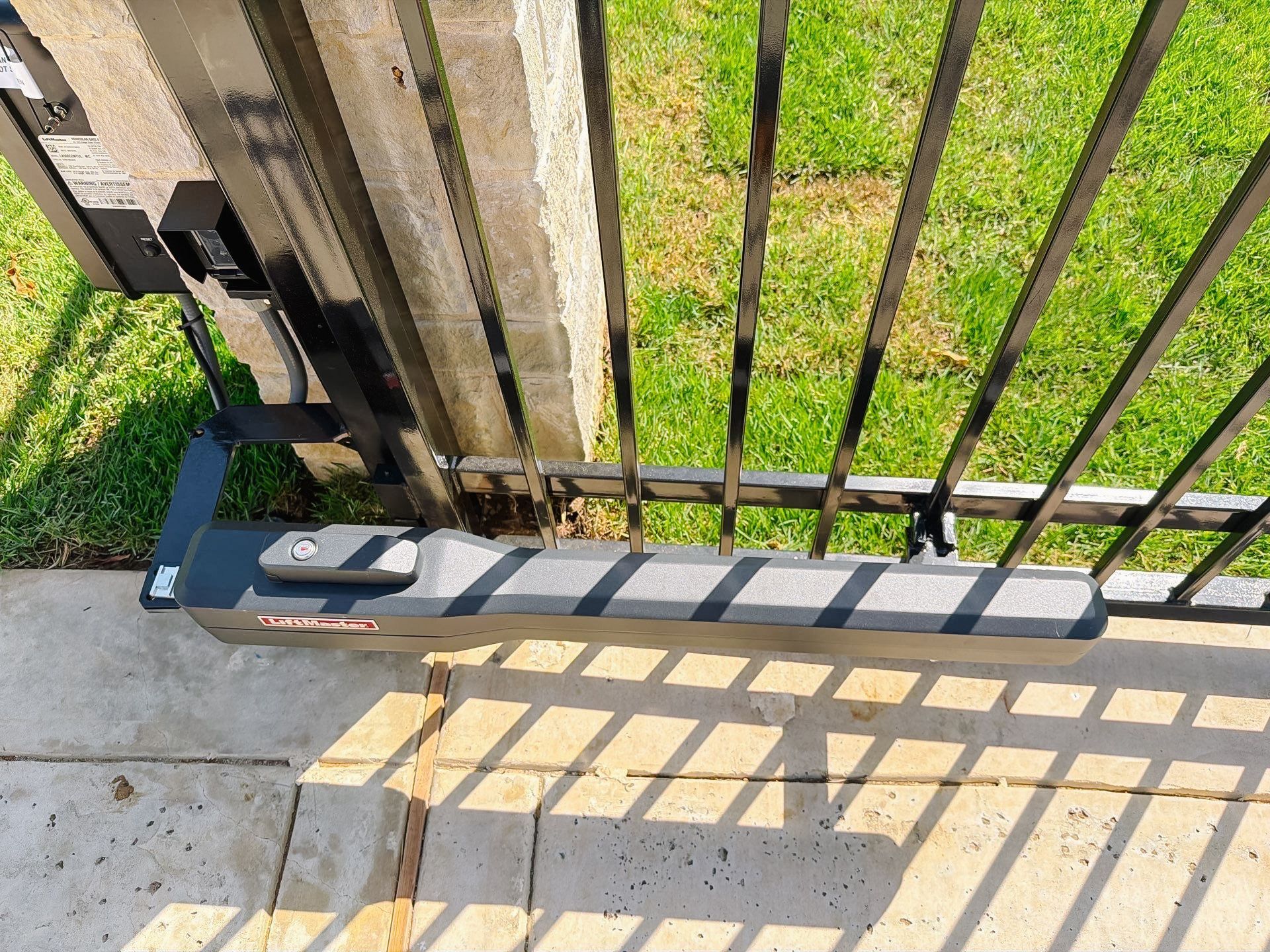 Black metal gate above concrete step, with sunlit grass and railing shadows on the ground