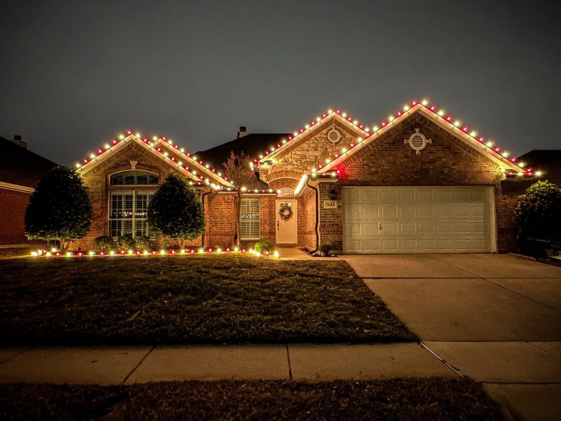 A brick house at night with Christmas lights outlining the roof and along the bushes.