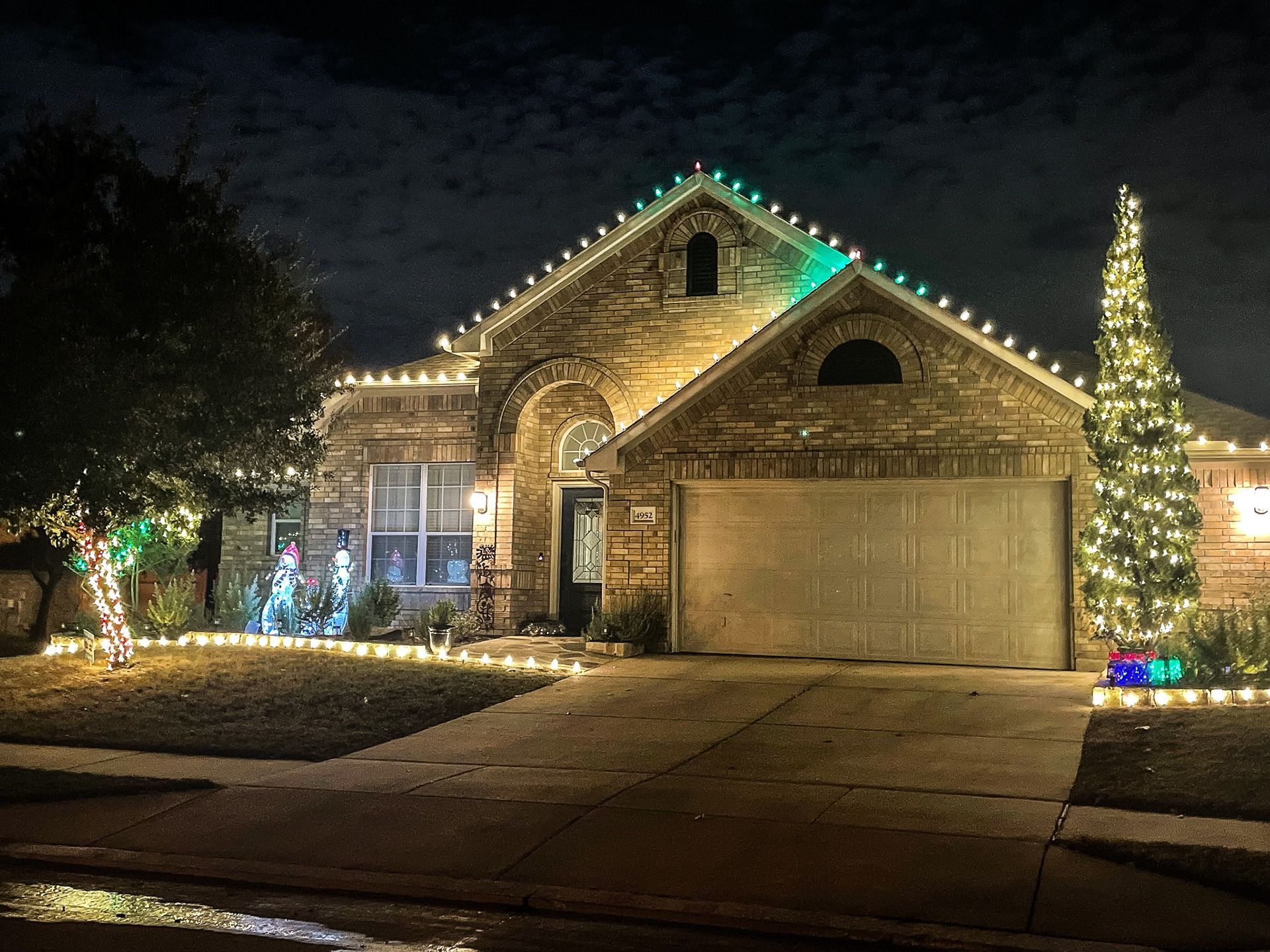 House decorated with Christmas lights, including the roofline, a tree, and landscaping.