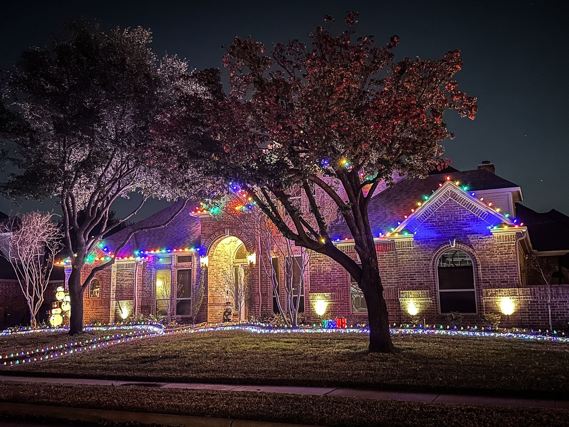 House decorated with colorful Christmas lights at night, illuminating trees and yard.