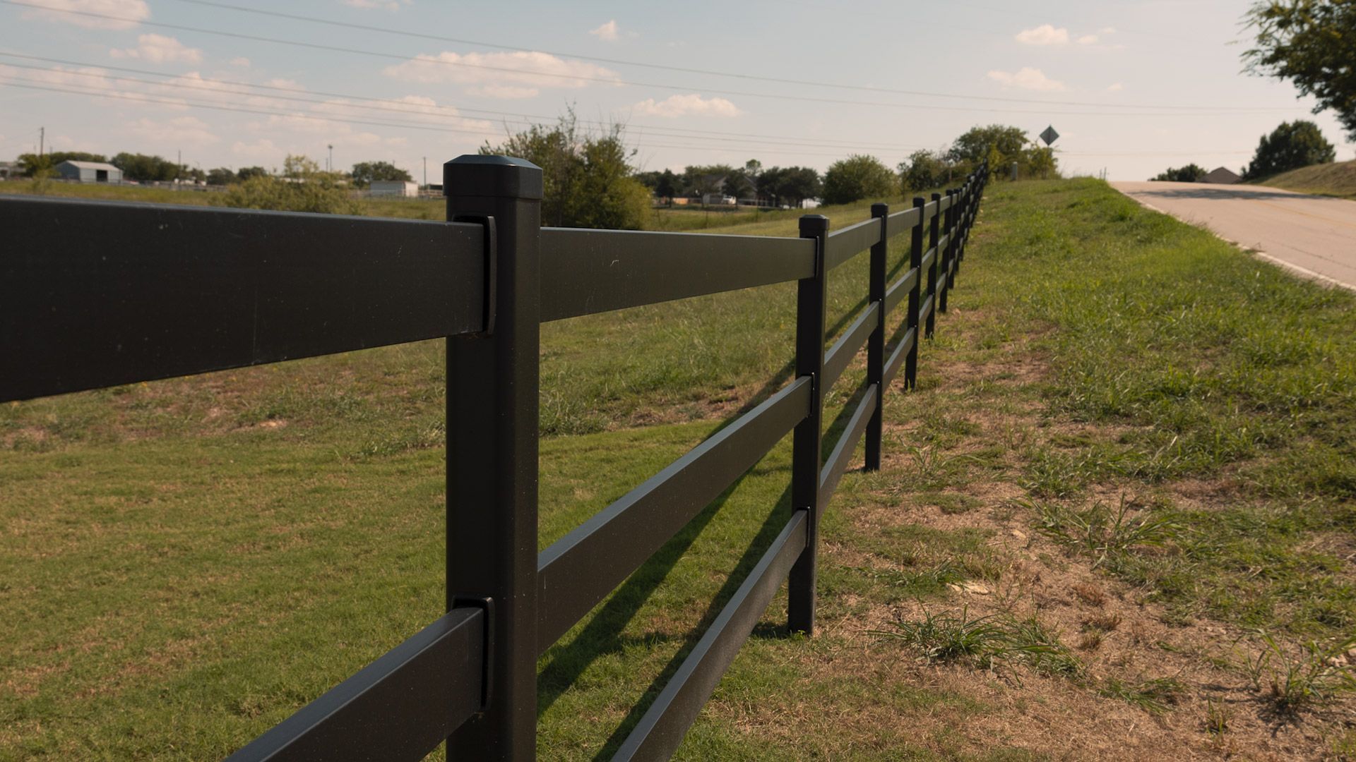 Black fence bordering a grassy area next to a road, under a sunny sky.