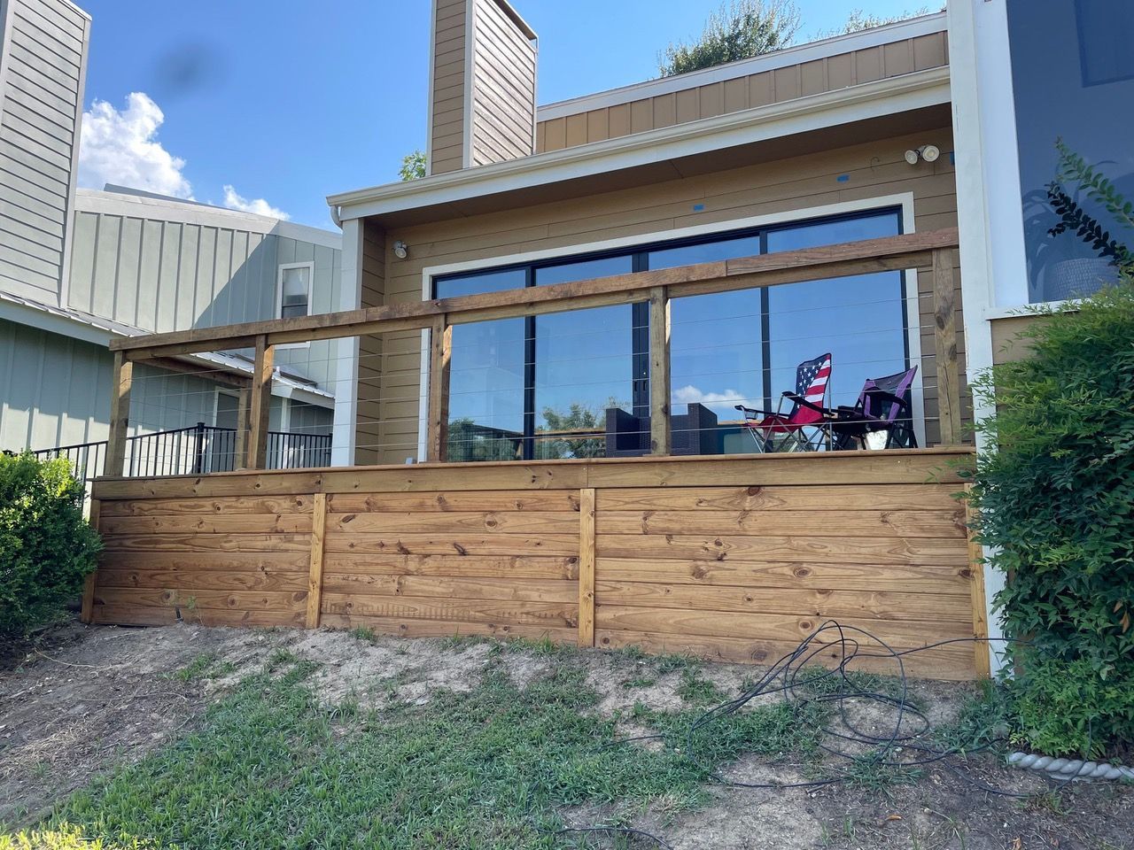 Wooden deck attached to a brick building with large windows. Lawn and foliage in the foreground; blue sky above.