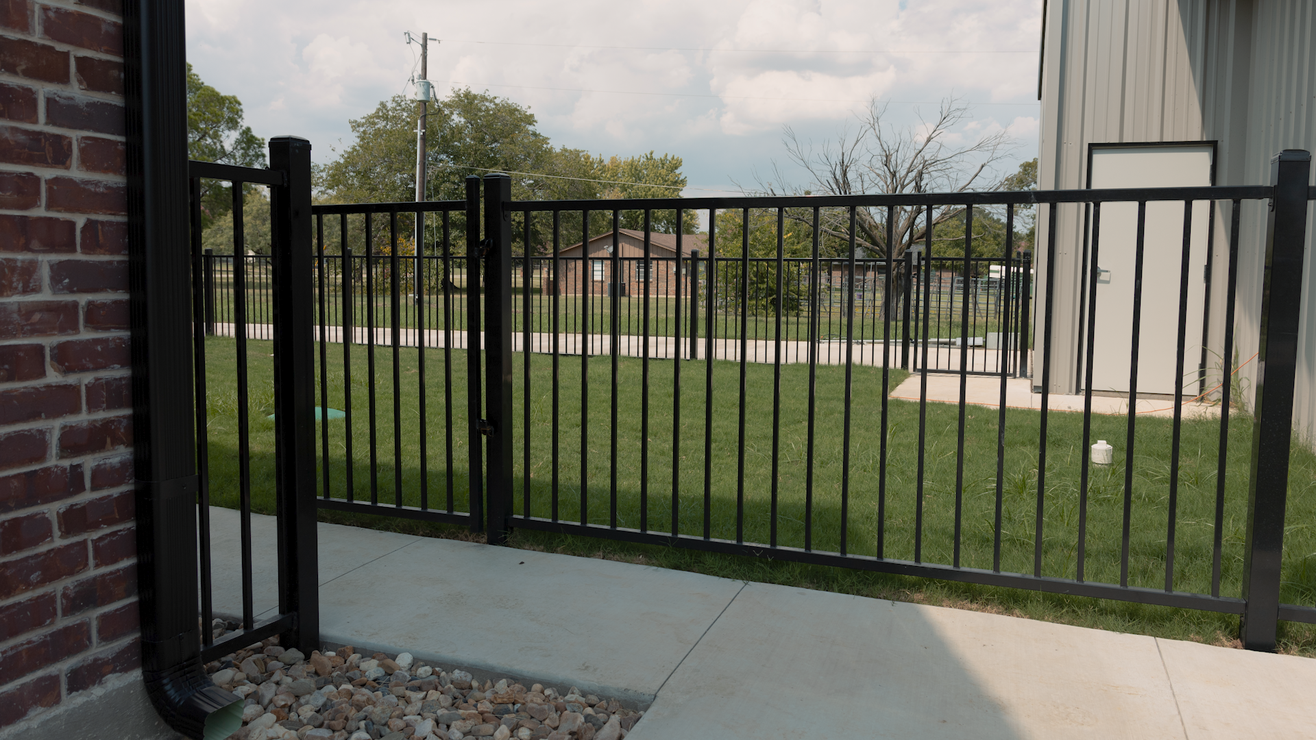 Black metal fence with gate, bordering a grassy area and building exterior, brick and concrete visible.
