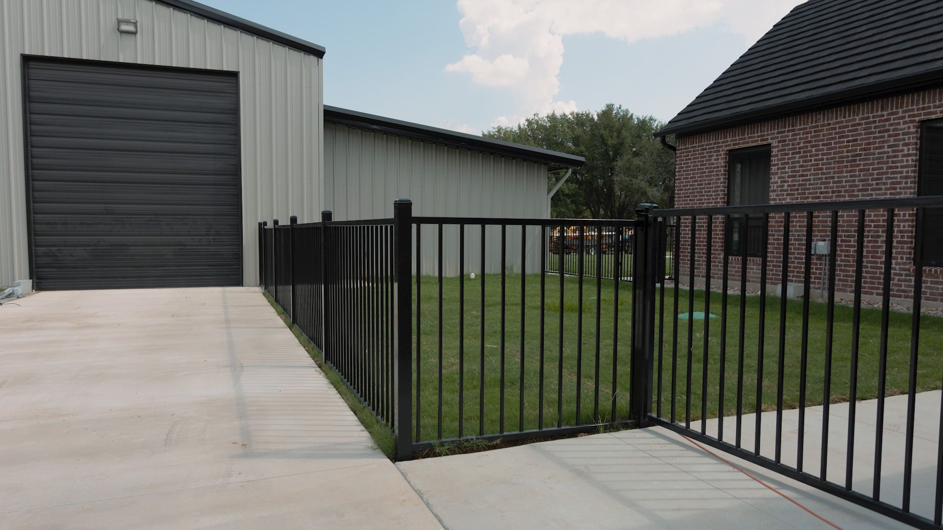 Black metal fence encloses a green lawn, next to a concrete driveway and two buildings with metal and brick exteriors.