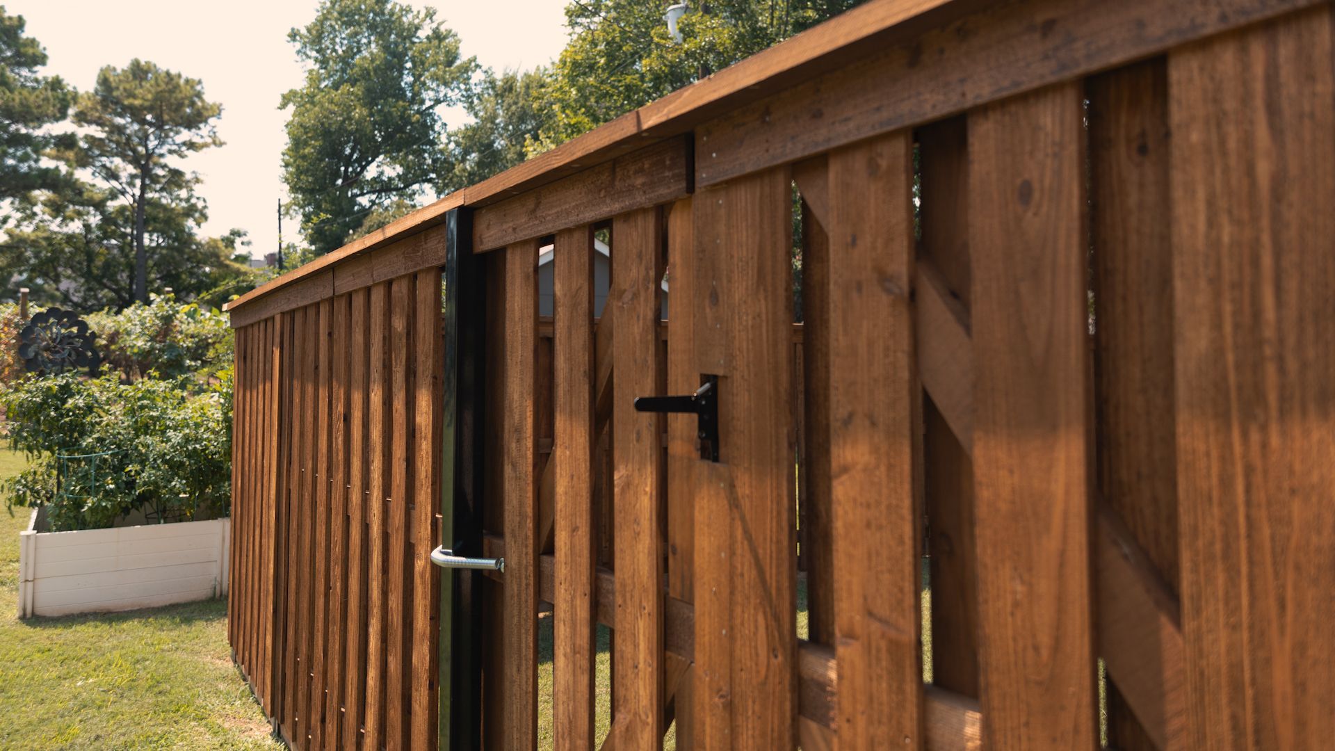 Wooden gate in a backyard, with black hardware. Tall, vertical boards; green trees in background.