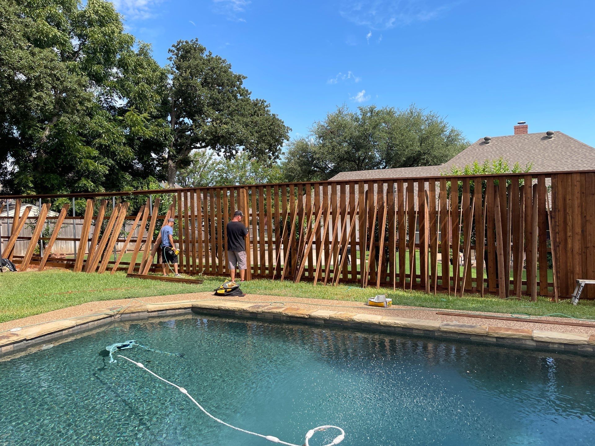 Two people are repairing a wooden fence along a backyard pool on a sunny day.
