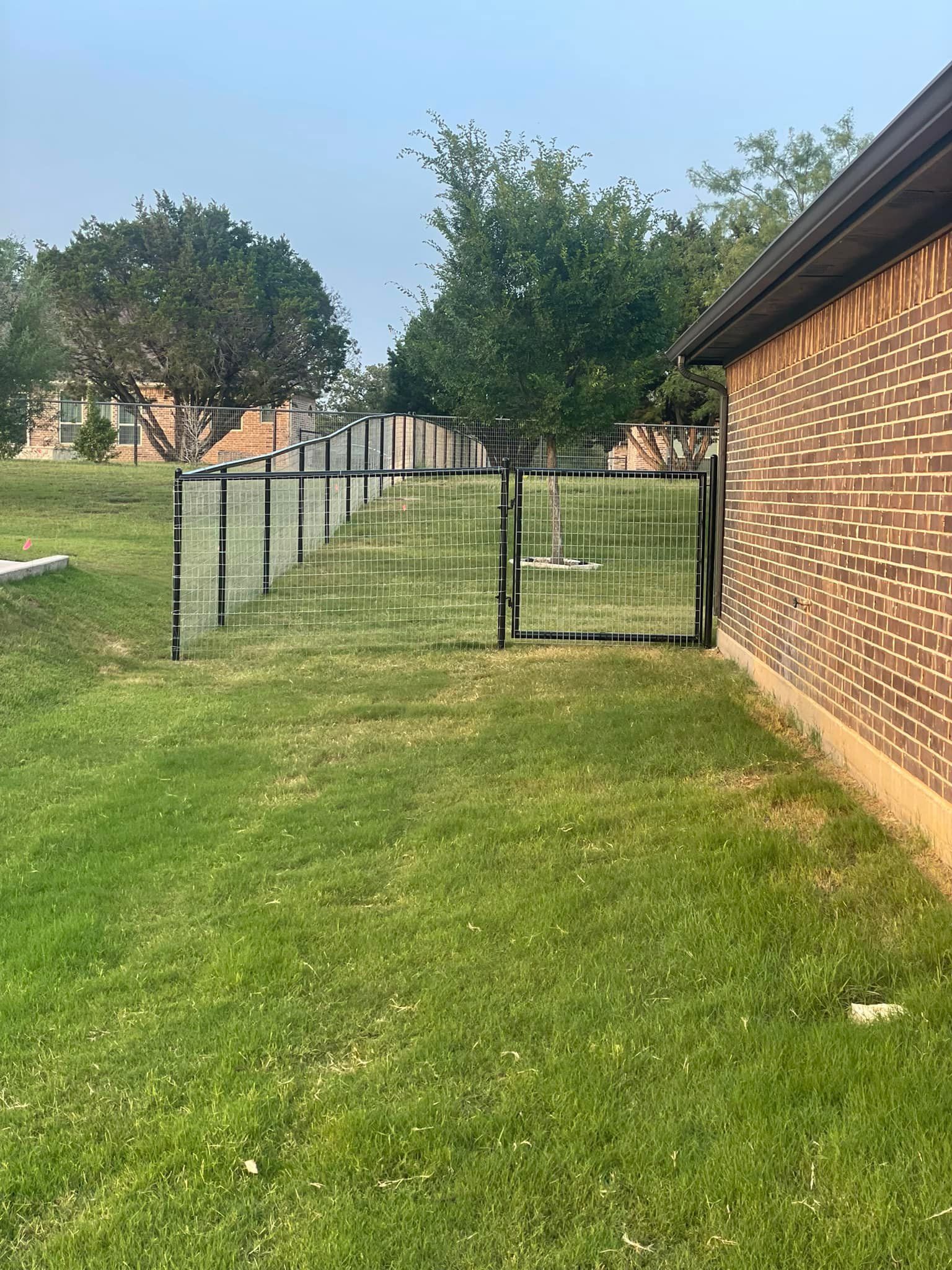 A fenced-in grassy yard next to a brick building. A gate is visible within the fence.