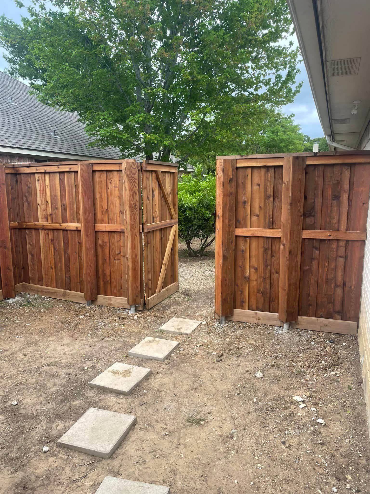 A wooden fence with an open gate, leading to a pathway of square stepping stones. The fence is brown, and the ground is dirt.